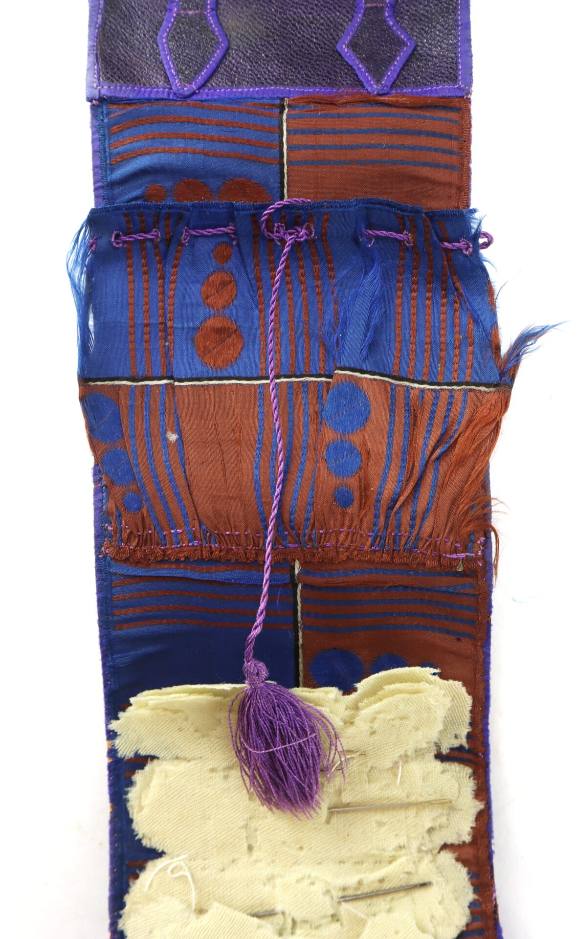 A textile piece with blue, brown, and cream sections, featuring geometric patterns, frayed edges, and a central purple cord with a tassel.