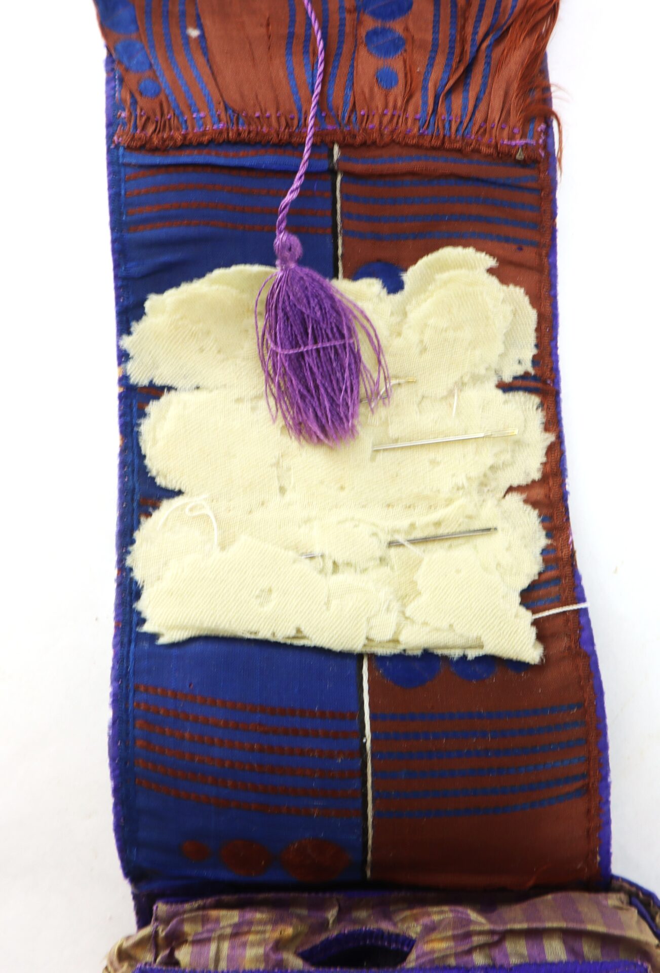 A fabric needle case with purple tassel, containing needles stuck in a padded, beige inner section, surrounded by patterned blue and brown fabric.