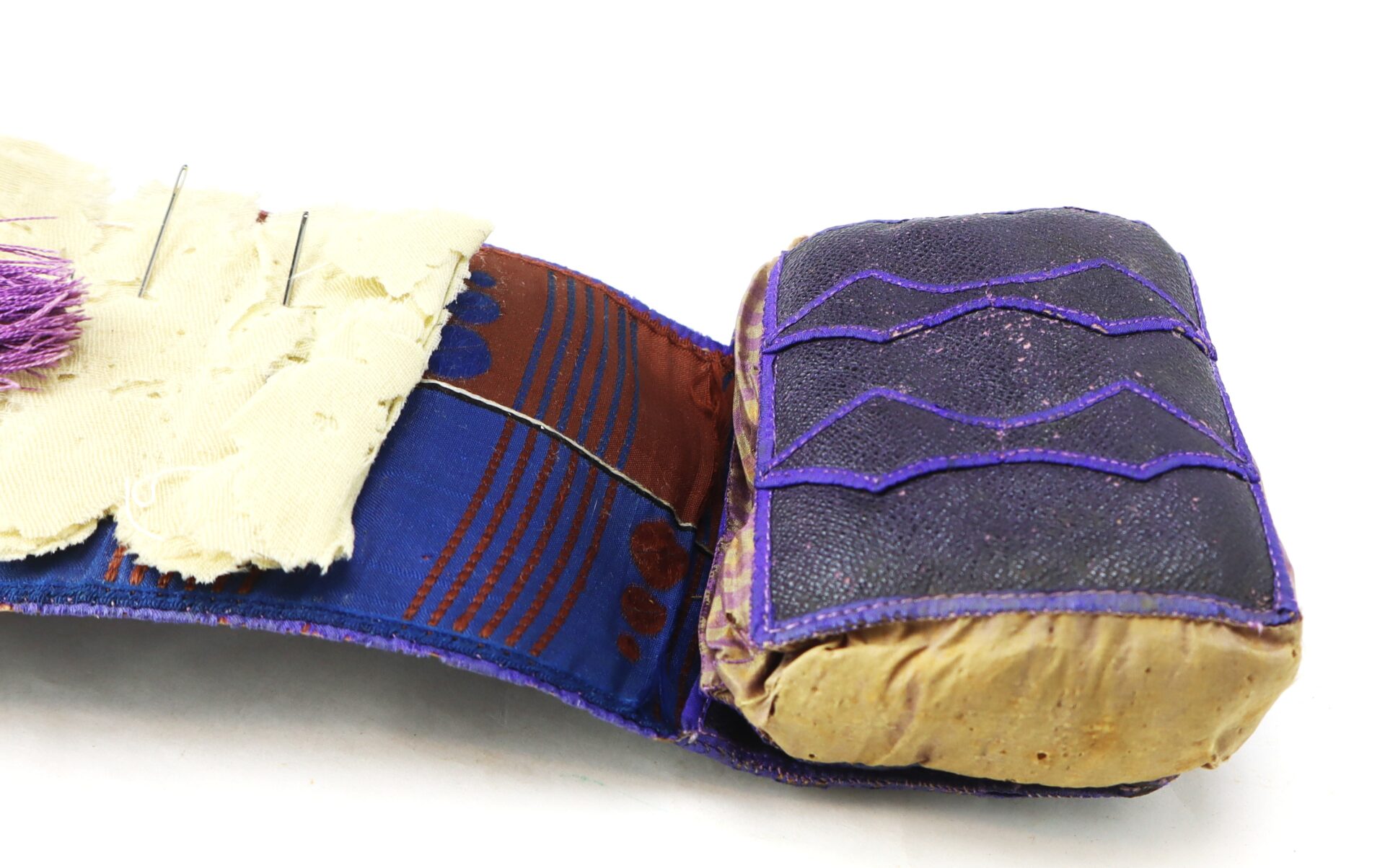 A close-up of a vintage purple and brown fabric pincushion with visible pins and stitching details.