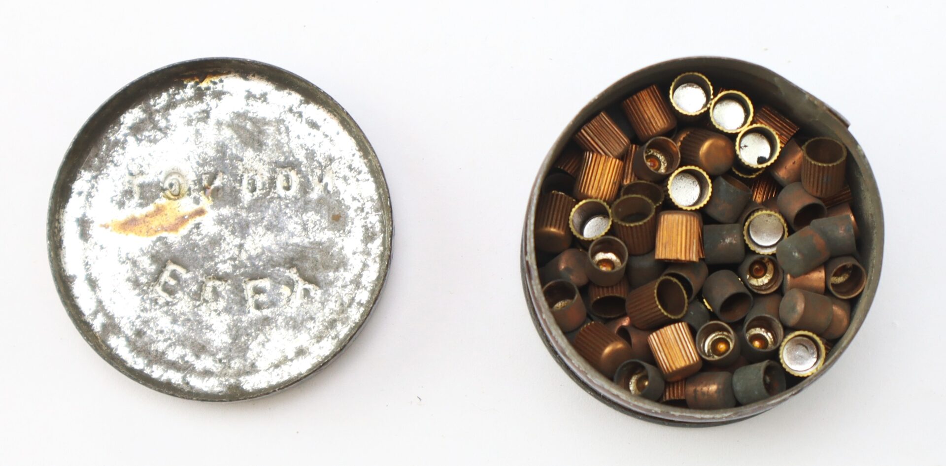 An open metal container with a lid and several small copper and gray percussion caps inside.