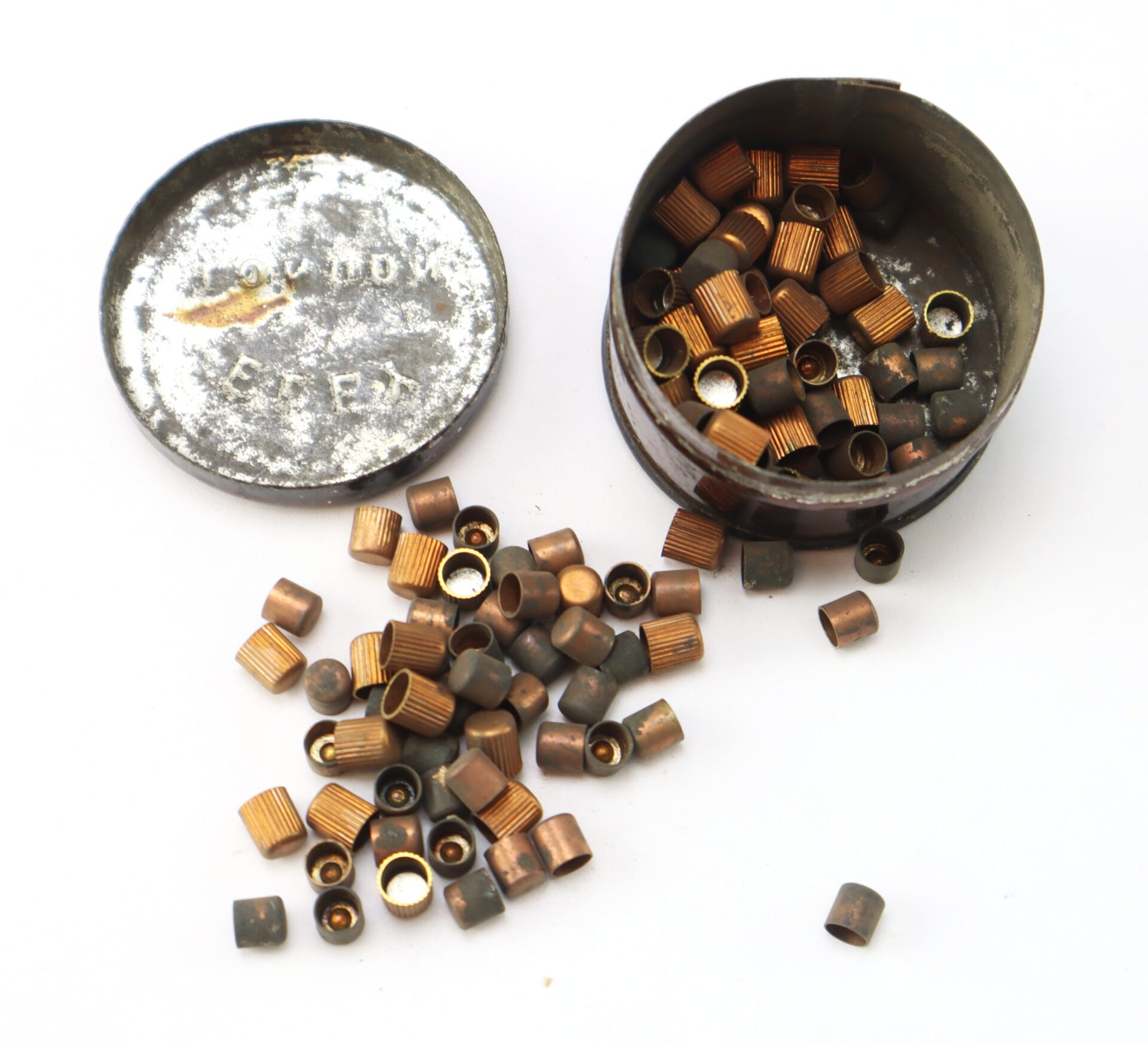 An open metal container with a detached lid, spilling out small copper-colored and gray percussion caps onto a white surface.