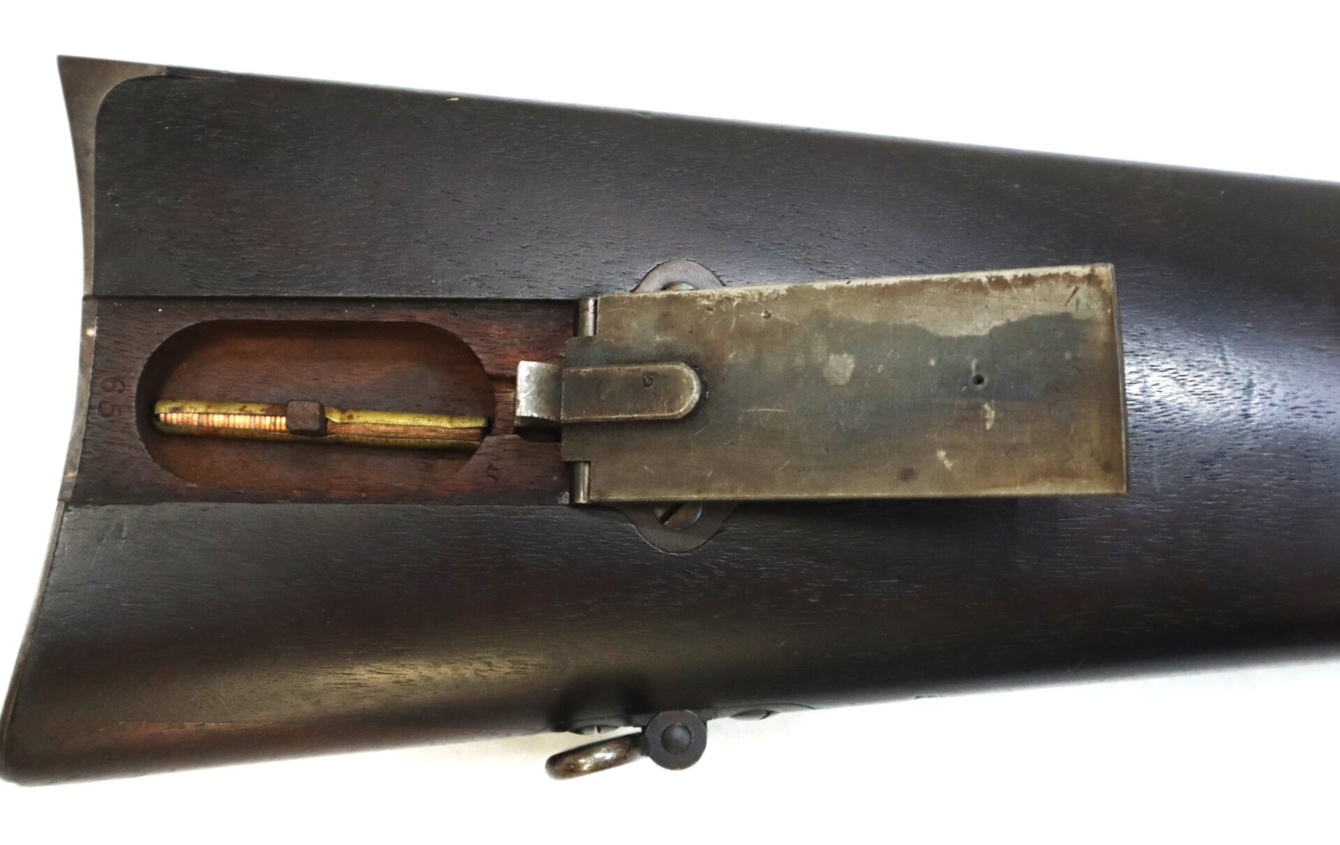Close-up of a rifle stock compartment with the metal cover open, revealing a cleaning rod and small storage space inside the wooden buttstock.