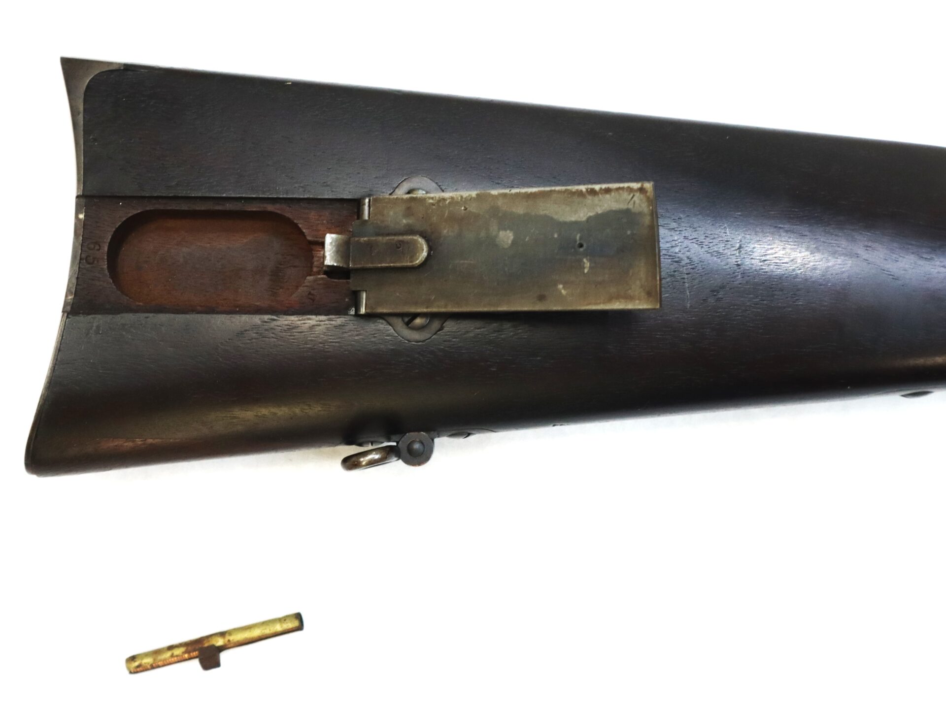 Close-up of a rifle stock with an open metal trapdoor compartment and a cleaning rod placed below it on a white background.