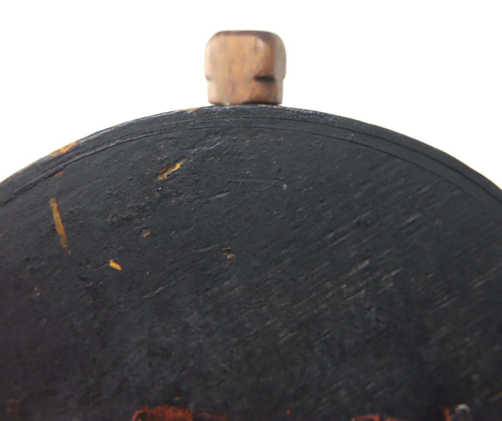 Close-up of a black, worn surface with scratches and a wooden knob on top, showing the Mexican War Cheesebox Canteen against a white background.