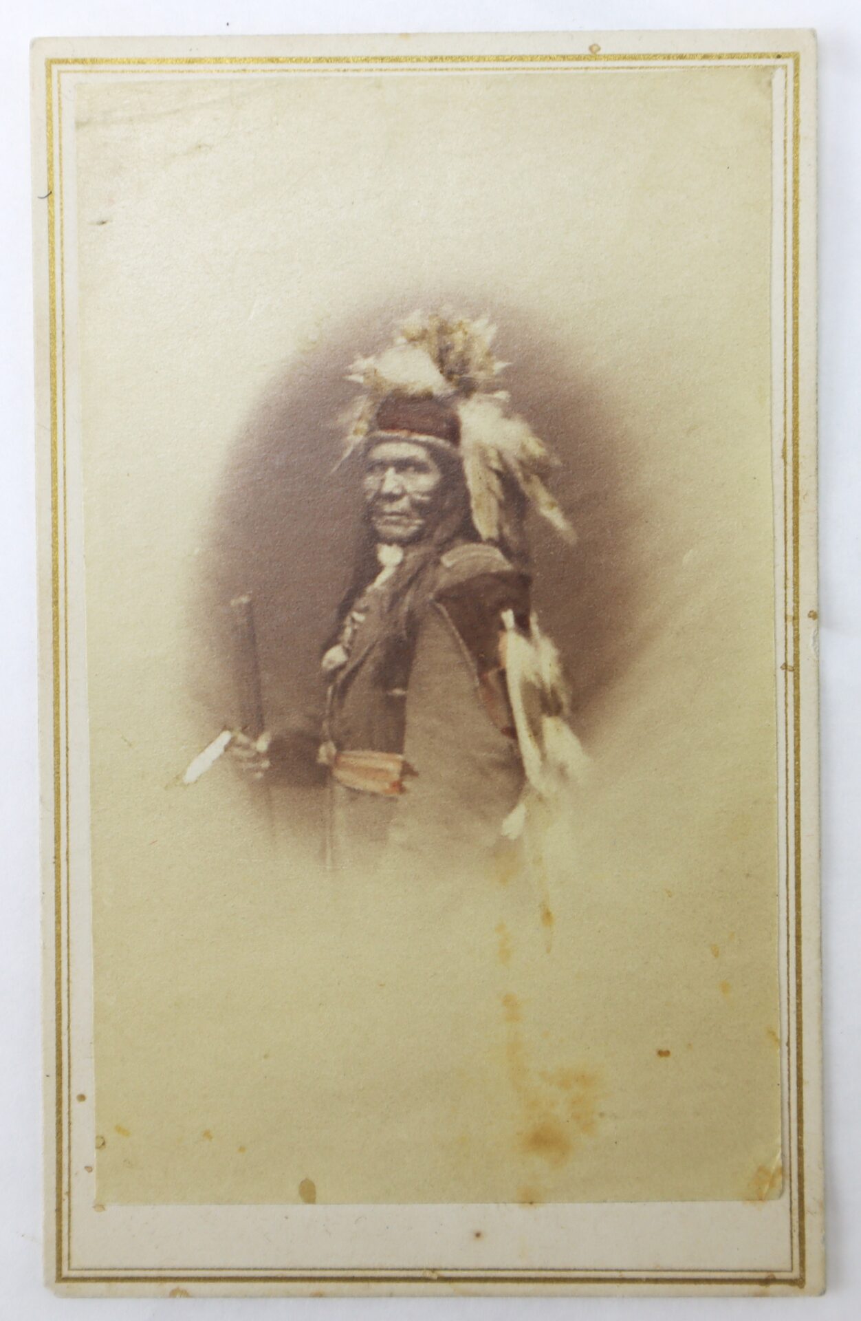 A sepia-toned portrait of a Native American man wearing traditional clothing and headdress, holding a staff, with a plain background and faded edges.
