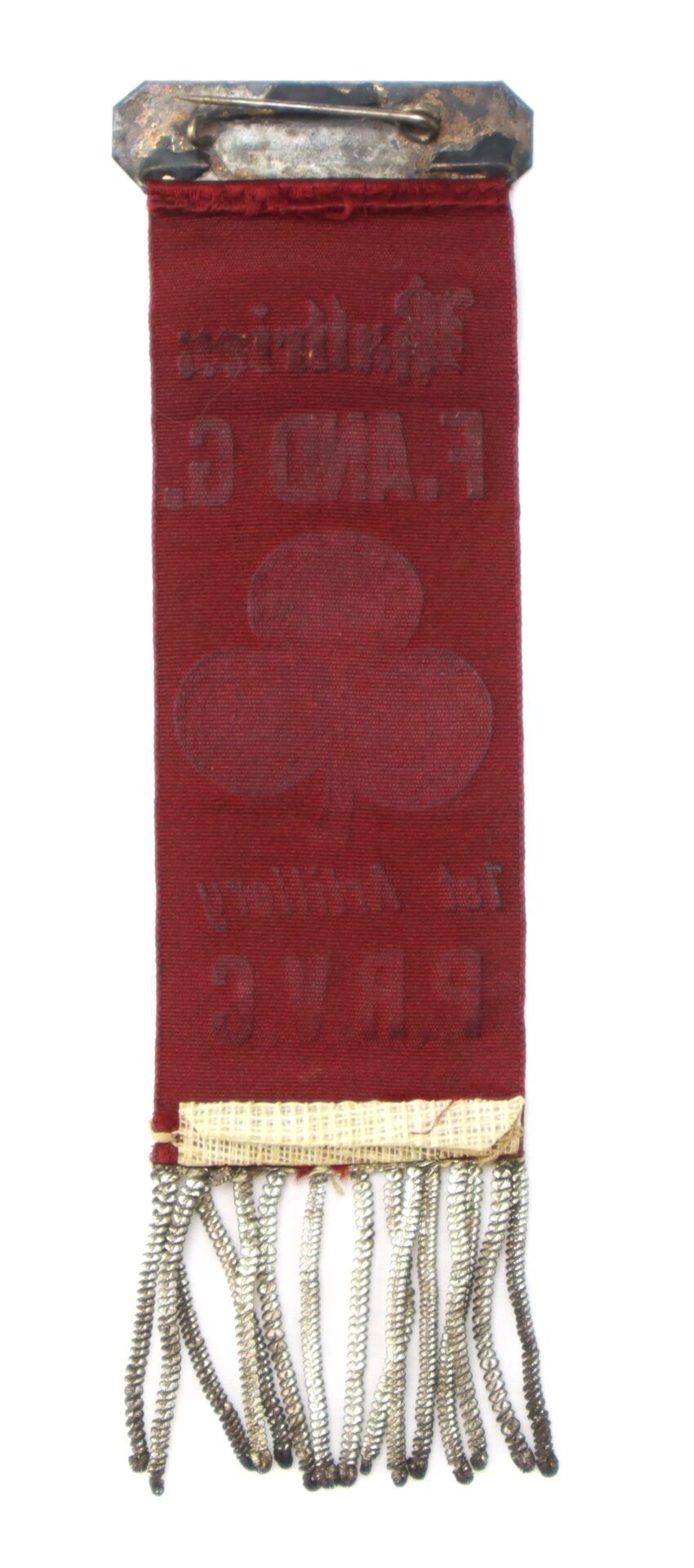 A vintage red ribbon badge with a metal clip, faded text, a clover emblem, and metallic fringe at the bottom.