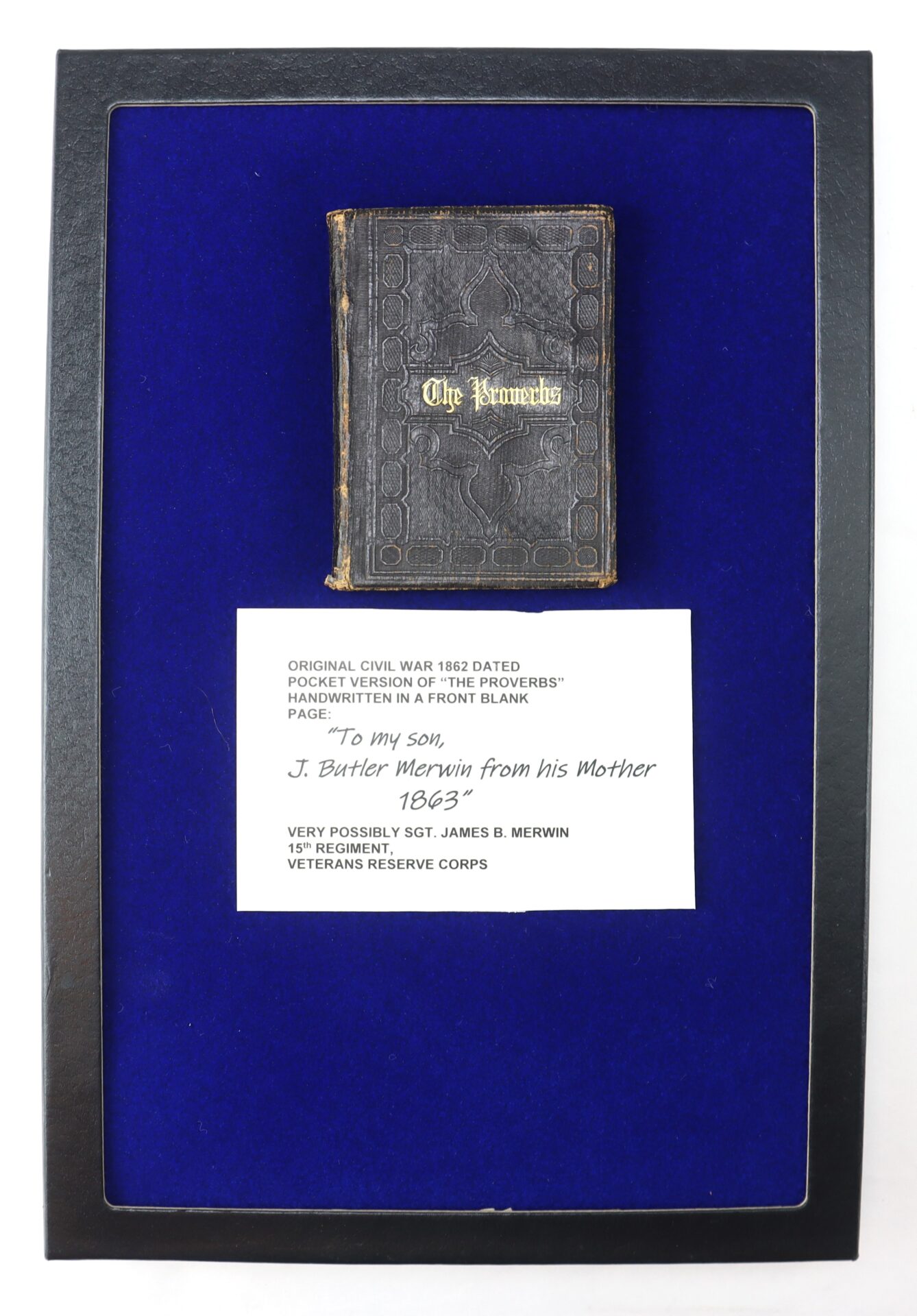 A small, worn book titled "The Proverbs" is displayed against blue fabric, accompanied by a label detailing its Civil War history and dedication from a mother to her son in 1863.