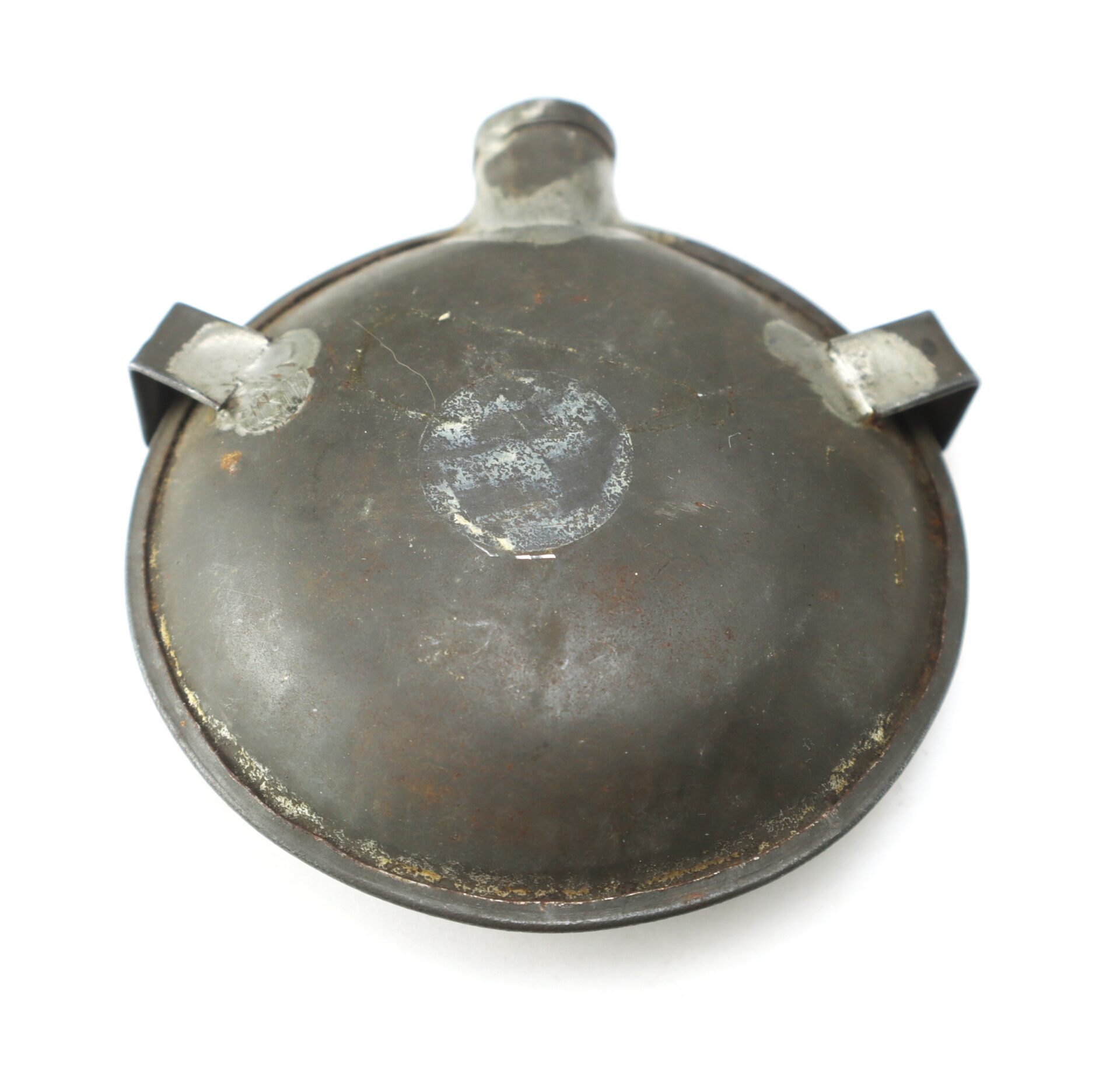 A round, metal military canteen cap with two small side brackets and a worn emblem in the center, showing signs of age and rust.