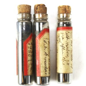Three small glass vials with cork stoppers and worn red-bordered labels, containing "Powder Taken from a Shell Shot in the Battle of Cold Harbor 50 years after the battle" by Pierre Burton - L. E. Groseclose.