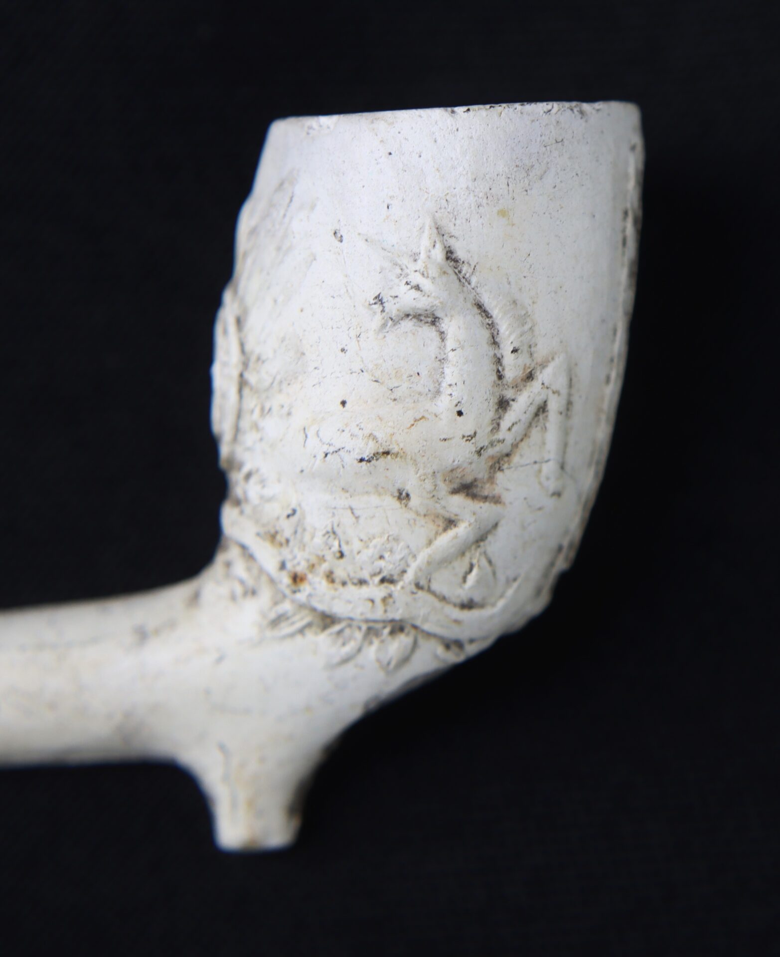 Close-up of a white clay tobacco pipe bowl with an embossed decorative pattern, set against a black background.