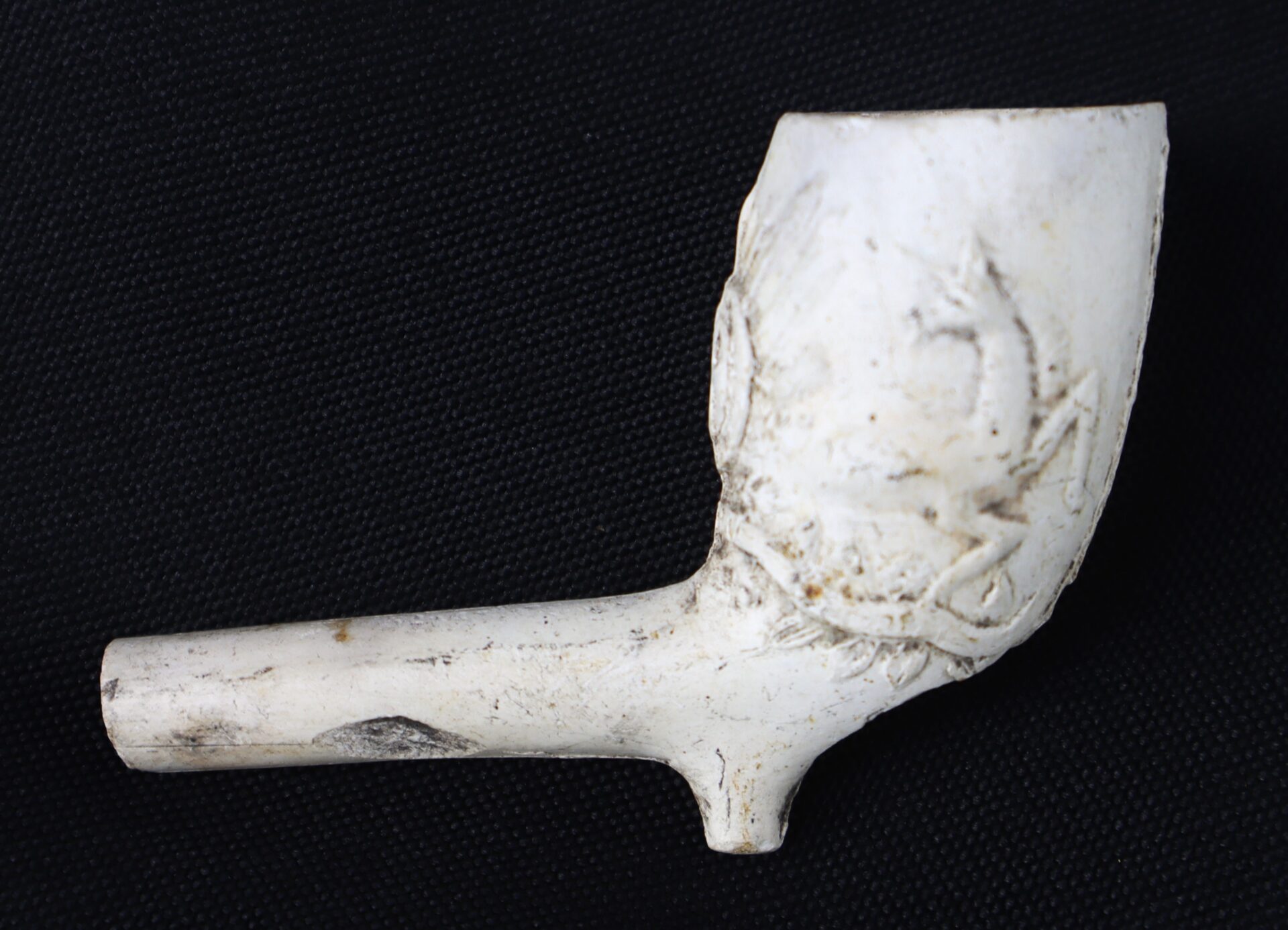 A white clay tobacco pipe with an embossed decorative design, shown against a black background.