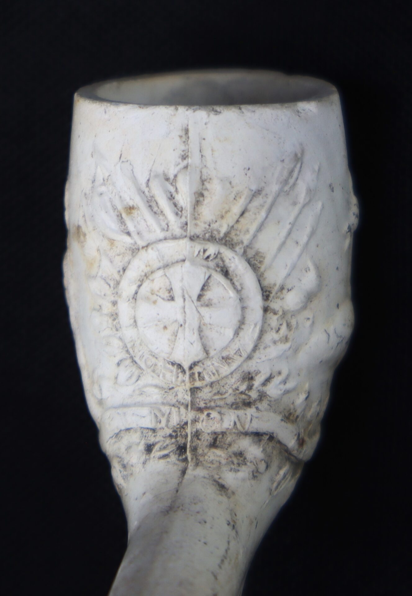 Close-up of a white clay pipe bowl featuring an embossed sunburst design and circular emblem with letters, set against a black background.
