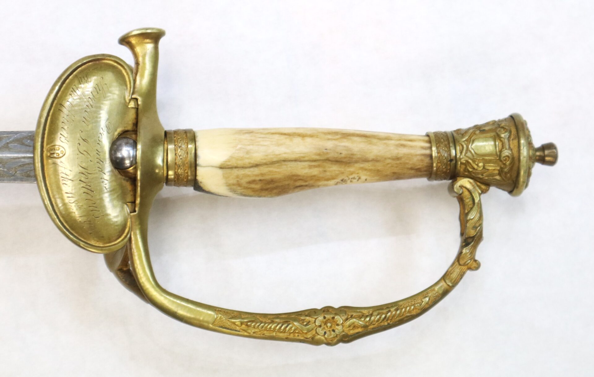 Close-up of an antique sword hilt featuring an ornate brass guard, an engraved knuckle bow, and a handle made of smooth, light-colored material.