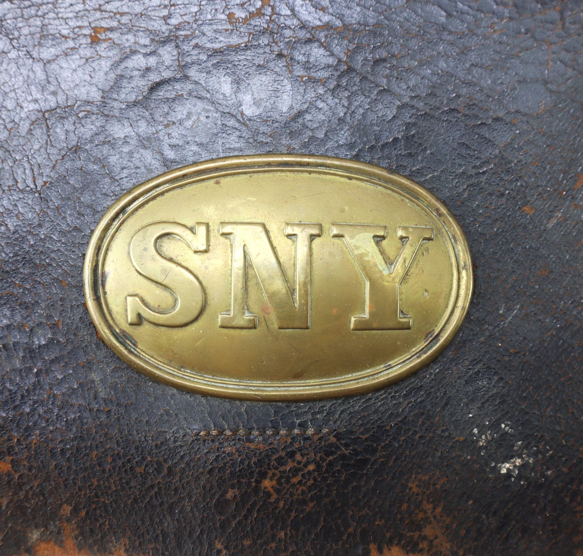 Oval brass plate with the letters "SNY" embossed on it, attached to worn, dark brown leather.
