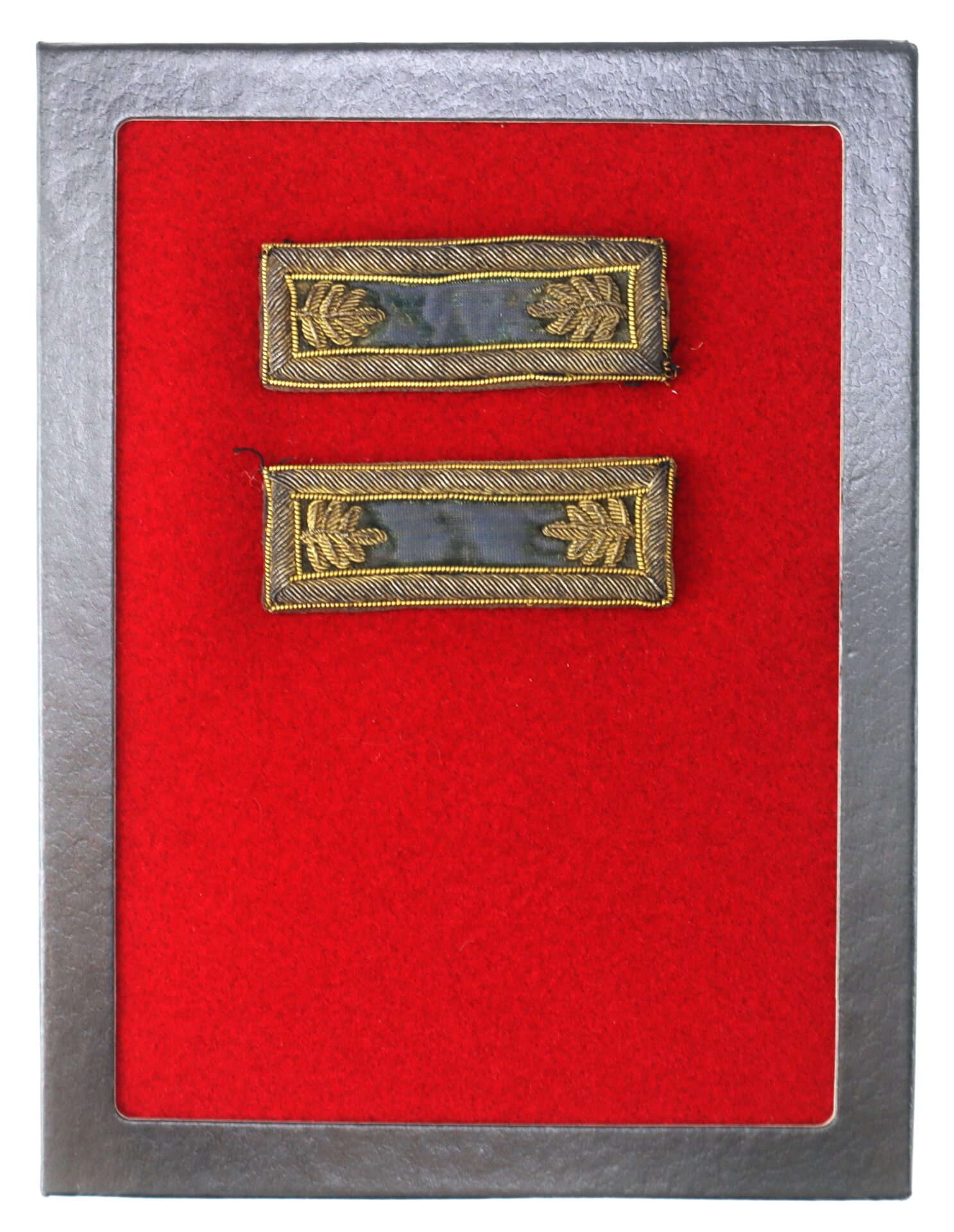 Two rectangular military rank insignia with gold embroidery displayed on a red felt background inside a metallic frame.