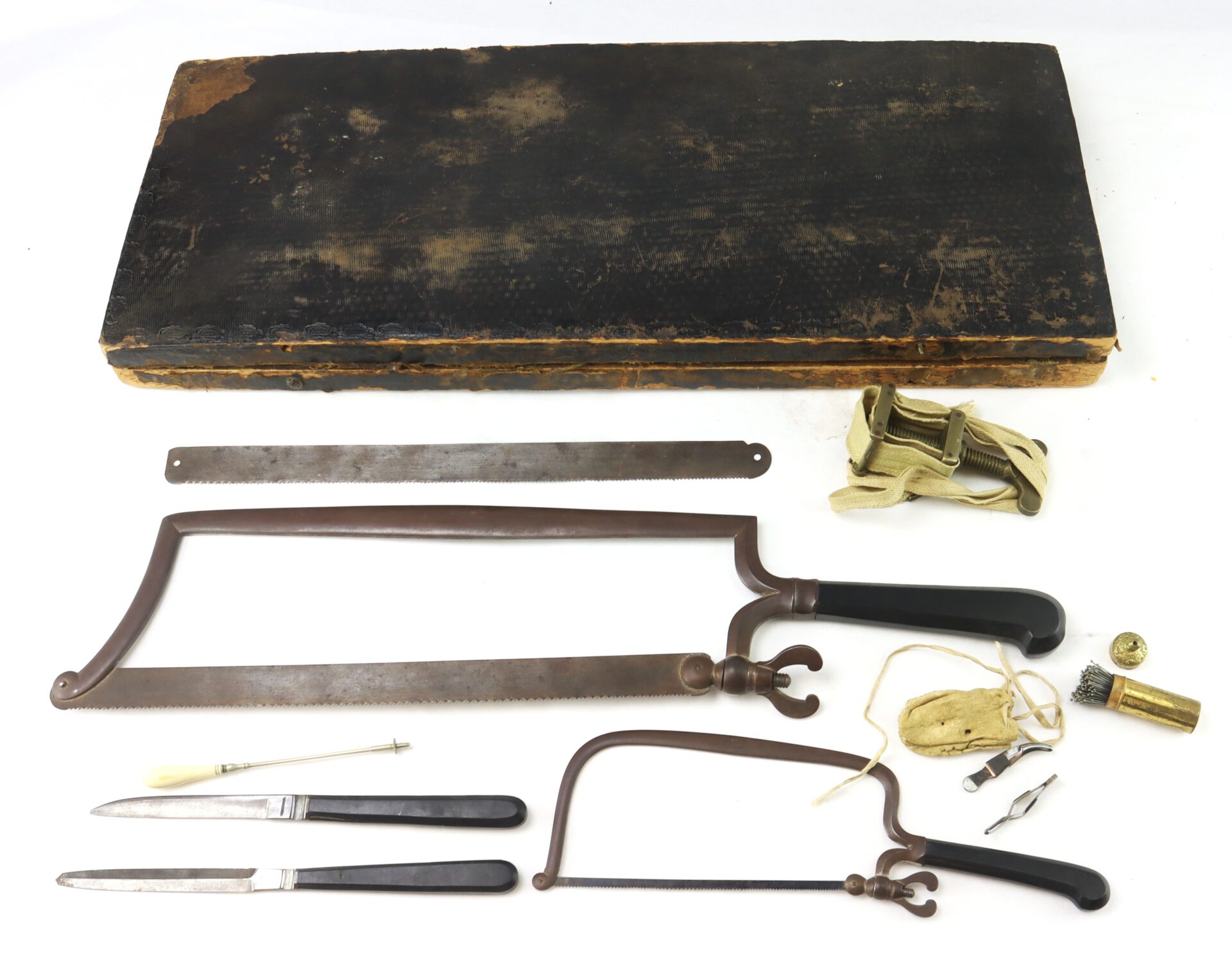 A set of vintage surgical tools, including saws, knives, scalpels, and other instruments, displayed below a worn black carrying case.