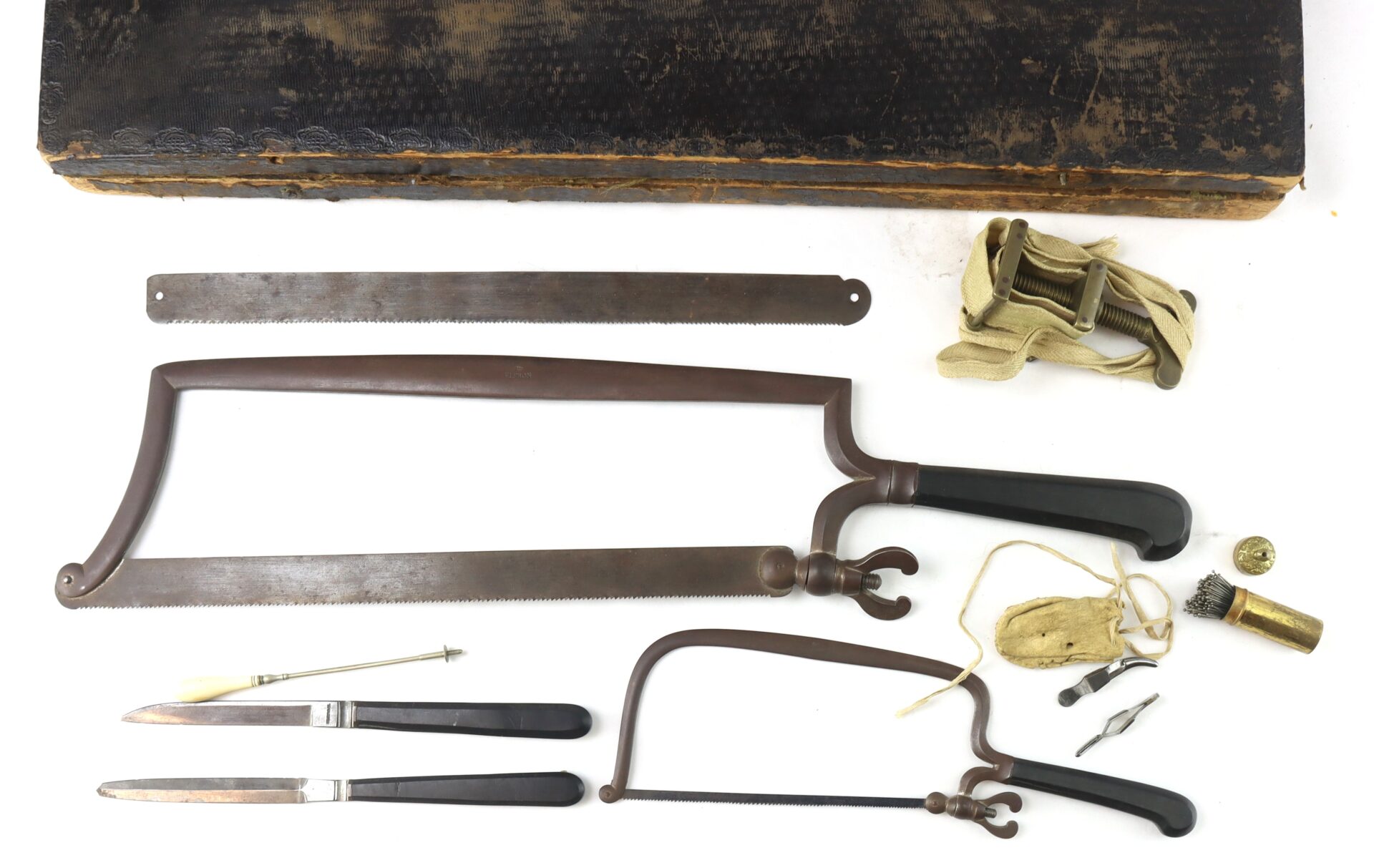 A vintage amputation kit with bone saws, knives, a tourniquet, a ruler, needles, thread, and other surgical tools displayed on a white surface.