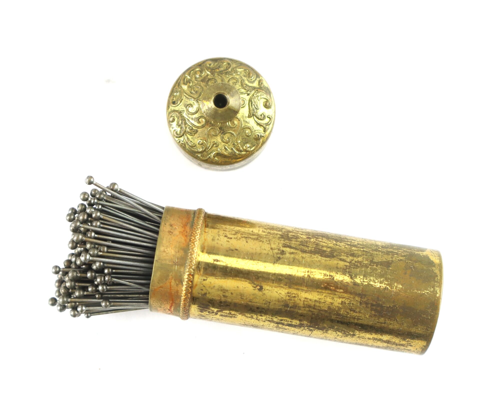A cylindrical brass container with a removable ornate lid holds multiple metal pins with rounded ends.
