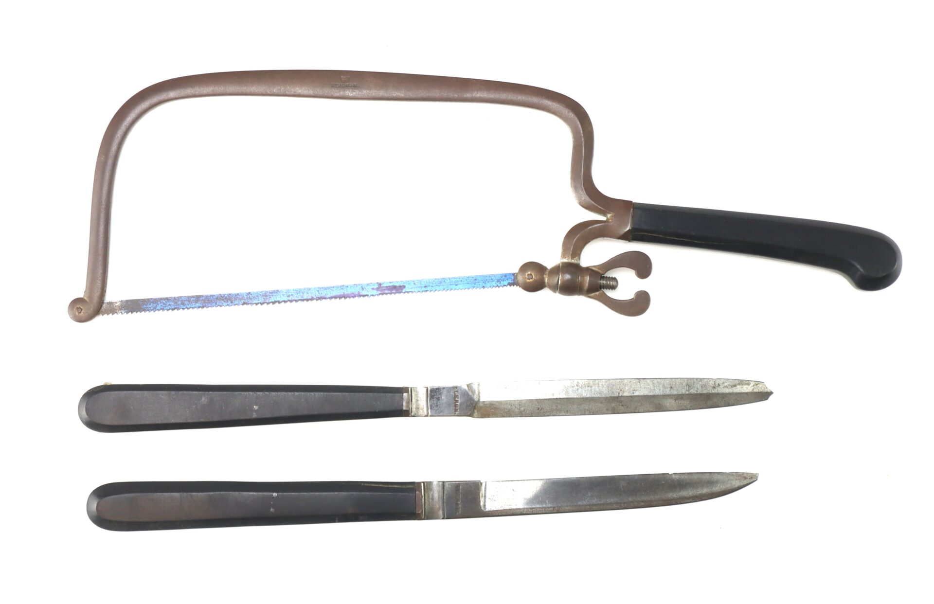 A bone saw with a metal frame and black handle is placed above two metal knives with black handles, all arranged against a white background.