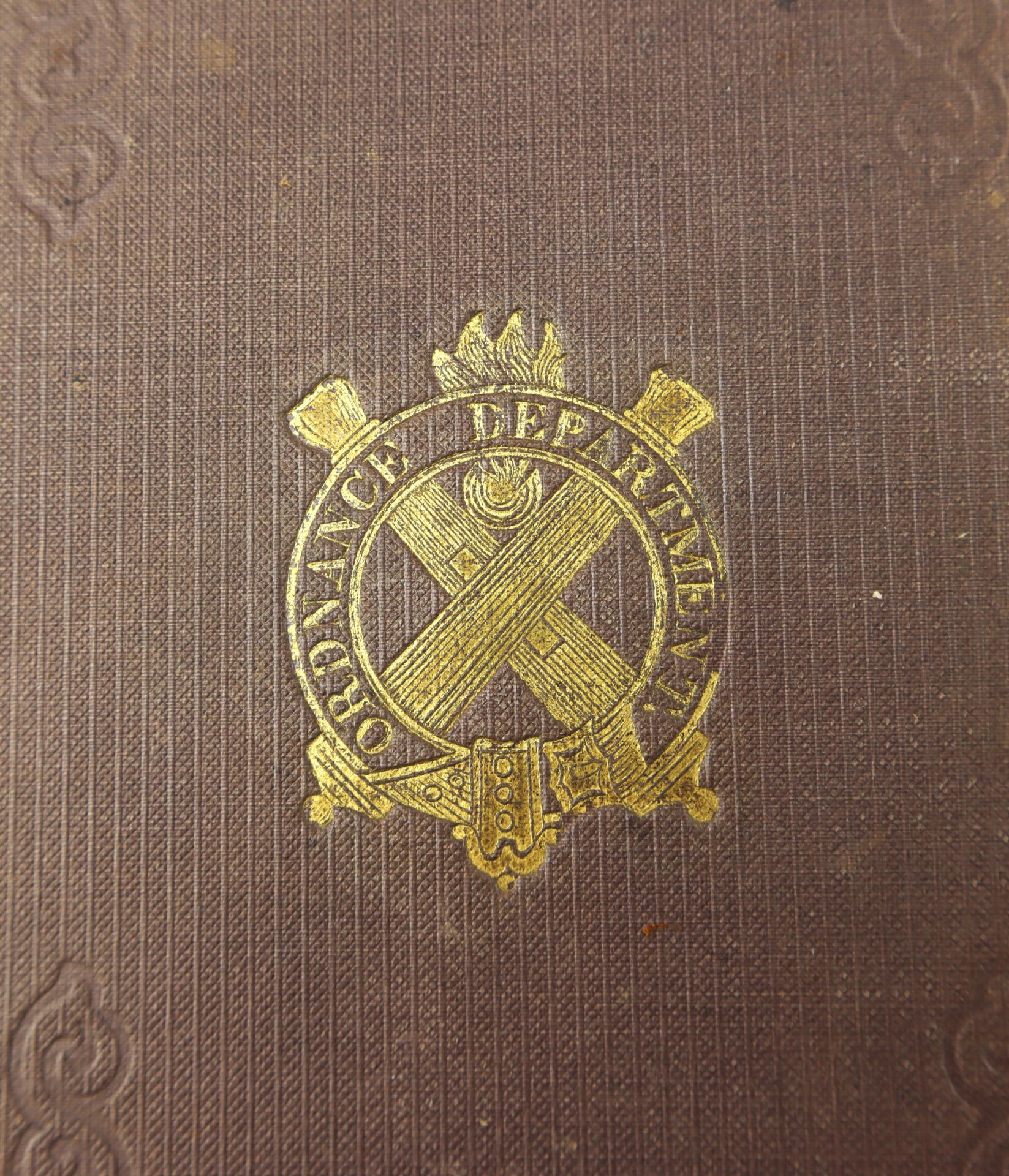 A close-up of the brown textured cover of the 1863 Ordnance Bureau Manual, identified to William A. Barker, 4th Maine Volunteer Infantry, featuring a gold-embossed "Ordnance Department" emblem with crossed cannons.