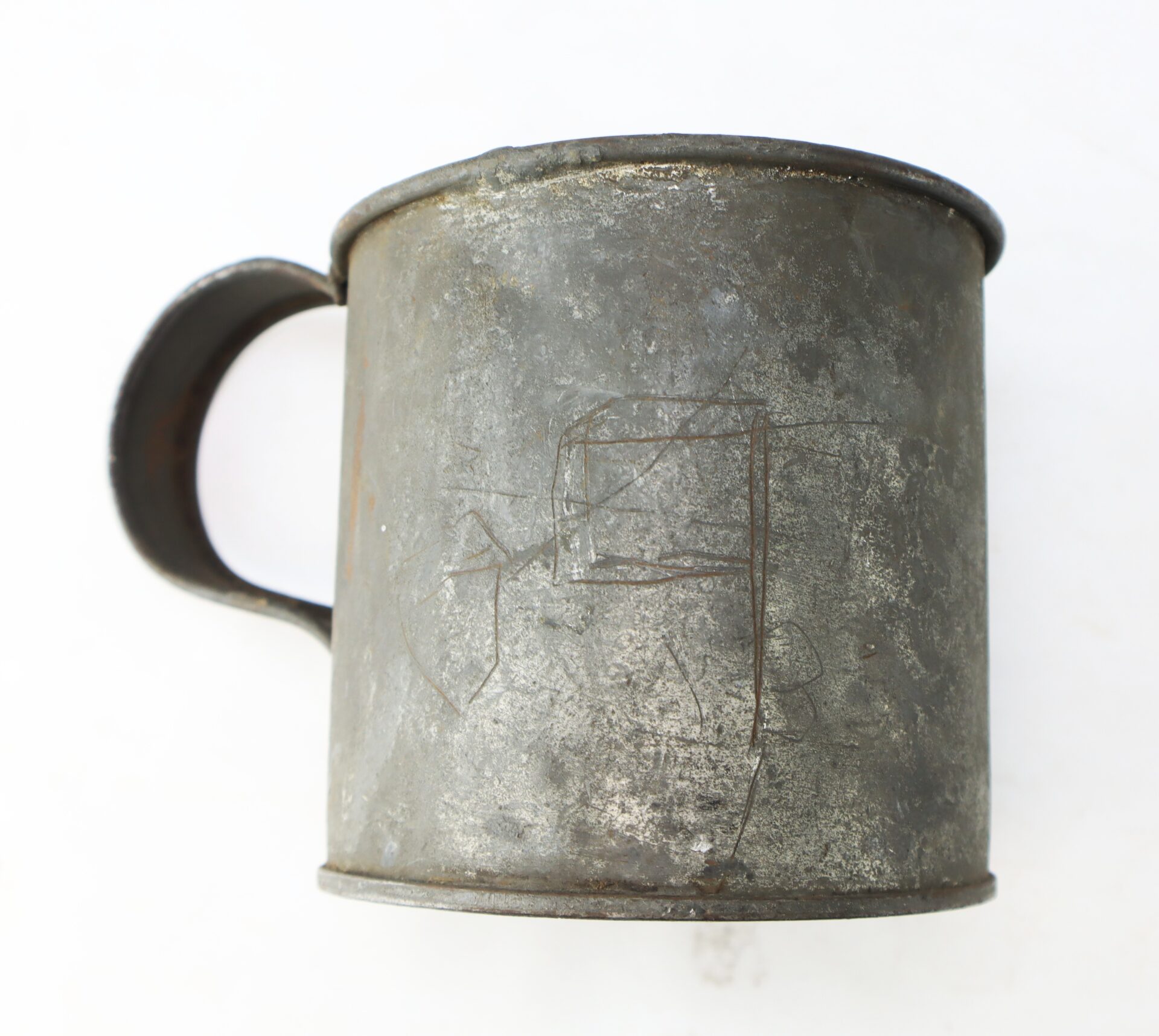 A weathered, metal cup with a handle and faint, scratched markings on its side, set against a plain white background.