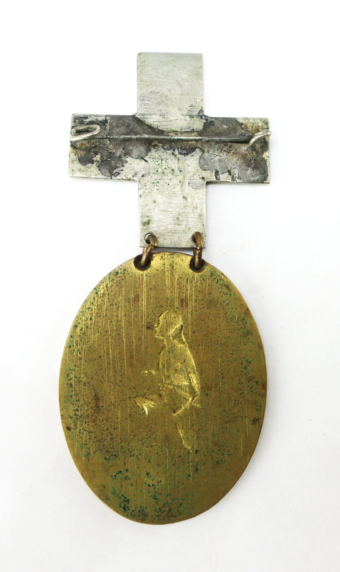 A tarnished, oval brass medal hangs from a roughly made metal cross; the medal features a faintly engraved figure in the center.