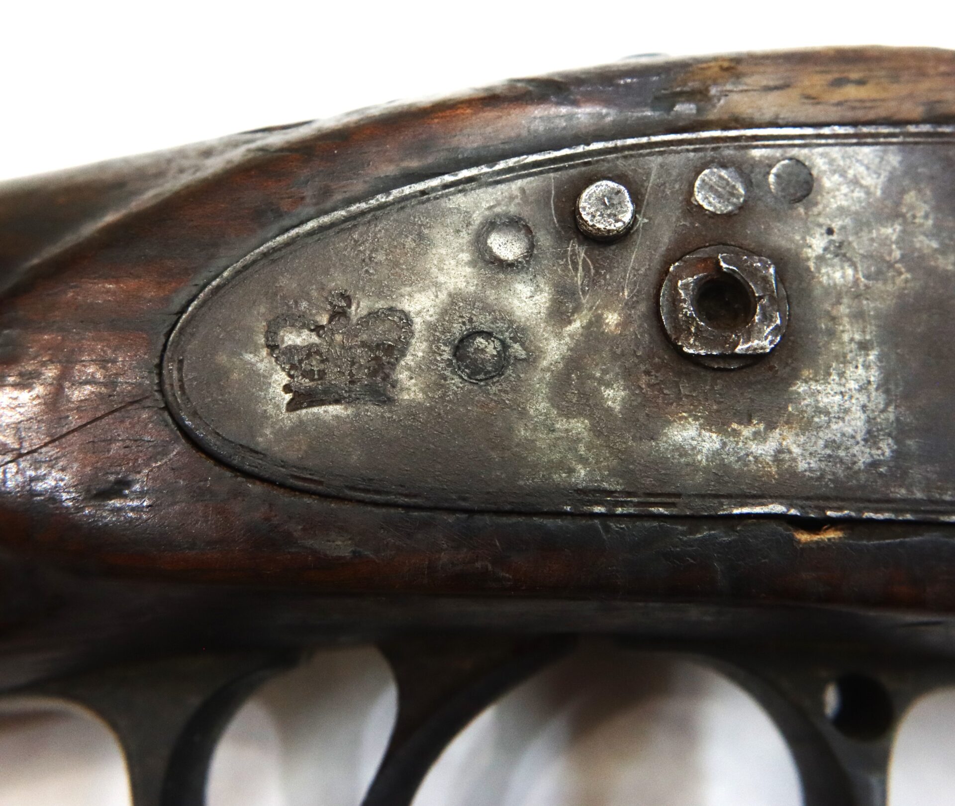 Close-up of a worn metal plate on a wooden firearm stock, showing stamped markings including a crown symbol and several dots above the trigger guard.