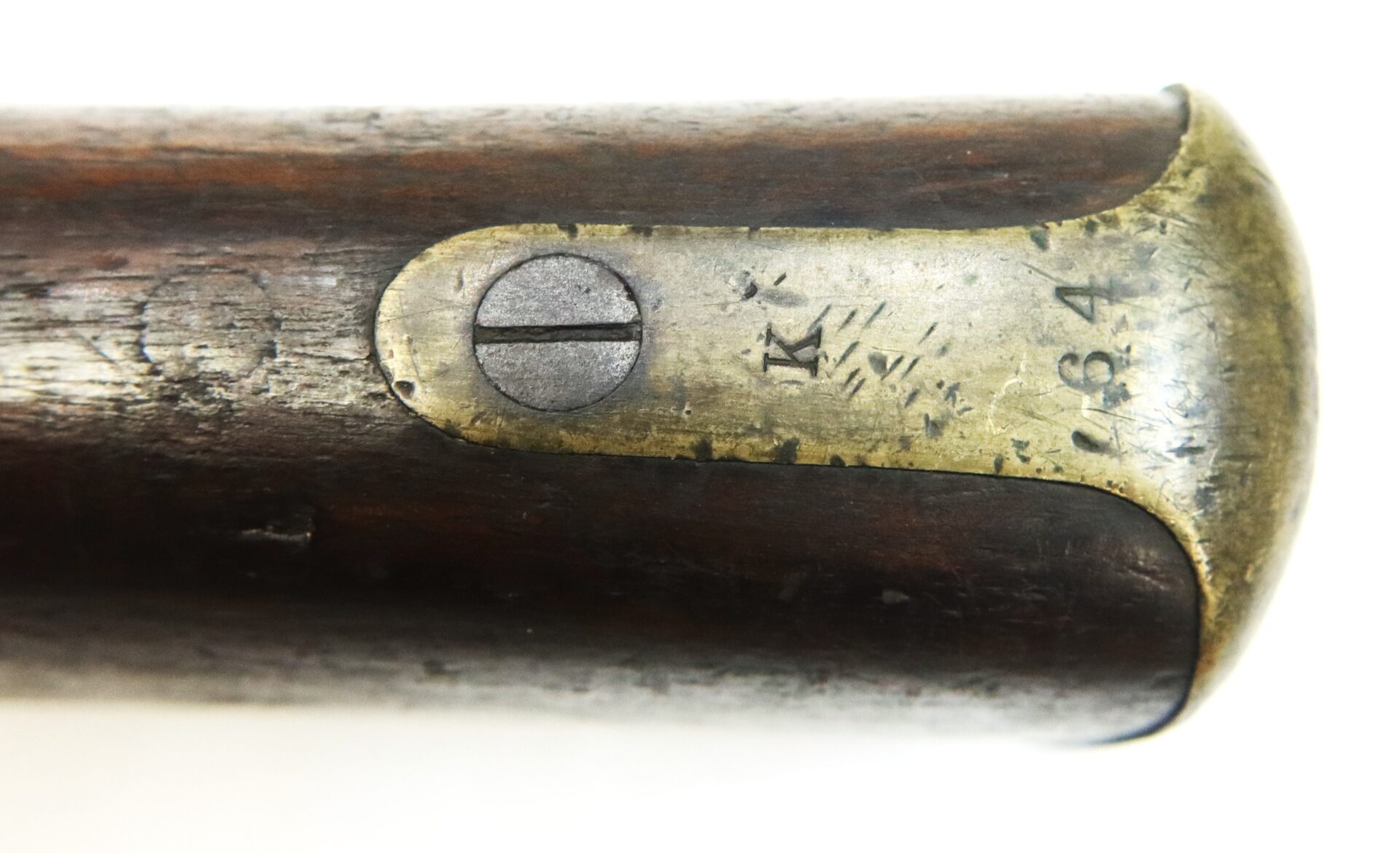 Close-up of a worn brass buttplate on a wooden rifle stock, featuring a flathead screw and engraved markings, including the number "84" and the letter "K.