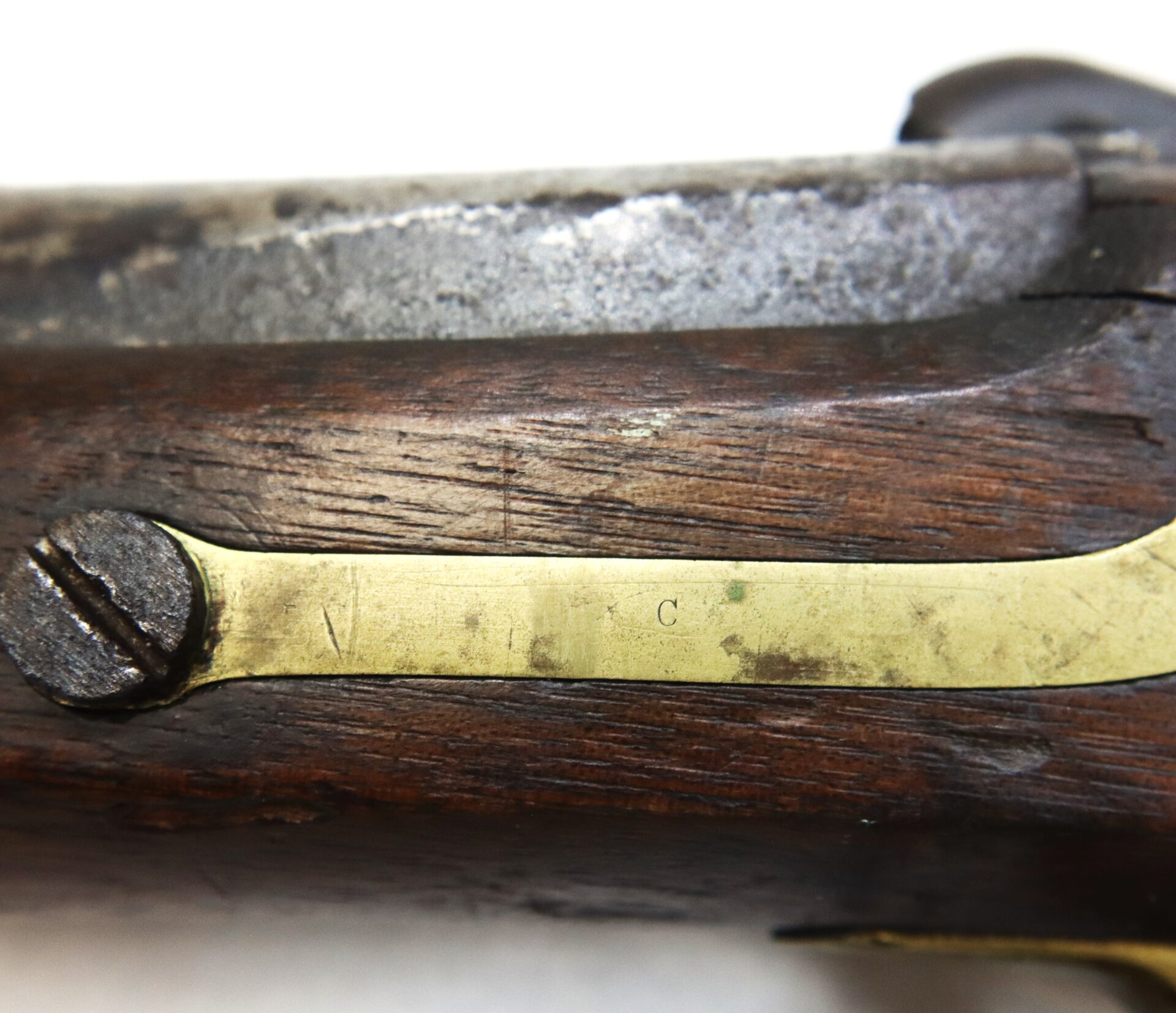 Close-up of an antique firearmās wooden stock, showing a brass plate with a screw and visible letter "C" engraved on the metal.