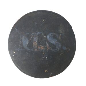A round, worn, black sign with faint, partially visible white letters "M.S." and two small holes near the bottom edge.