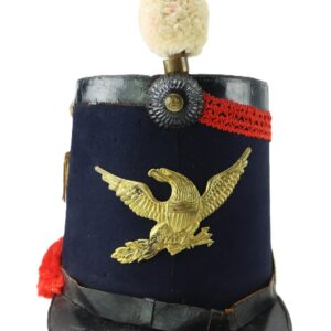 Shako military hat with a brass eagle emblem, dark blue fabric, black leather visor, red pompom and trim, and silver rosette at the front.