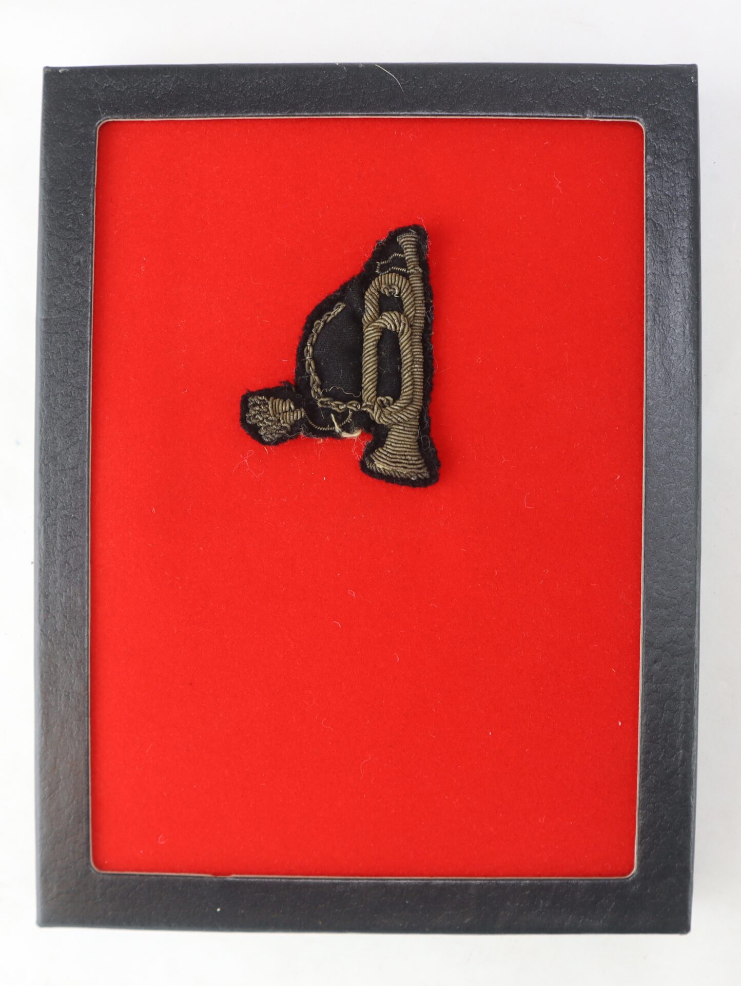 The Officer’s “Rifleman” Insignia: a small, embroidered military badge with a bugle, showcased in a black frame and set against a striking red background.