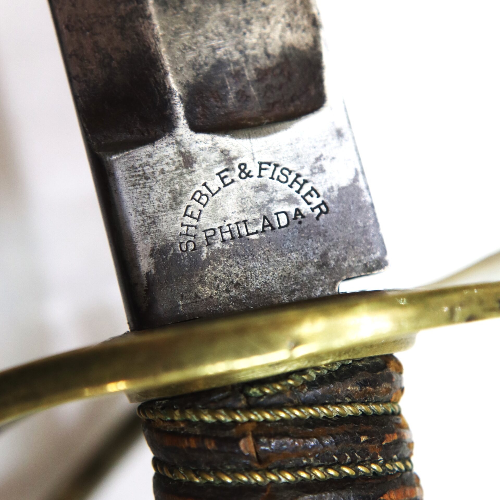 Close-up of a sword hilt showing engraved text "SHEBLE & FISHER PHILADA" on the metal near a brass guard and wrapped handle.