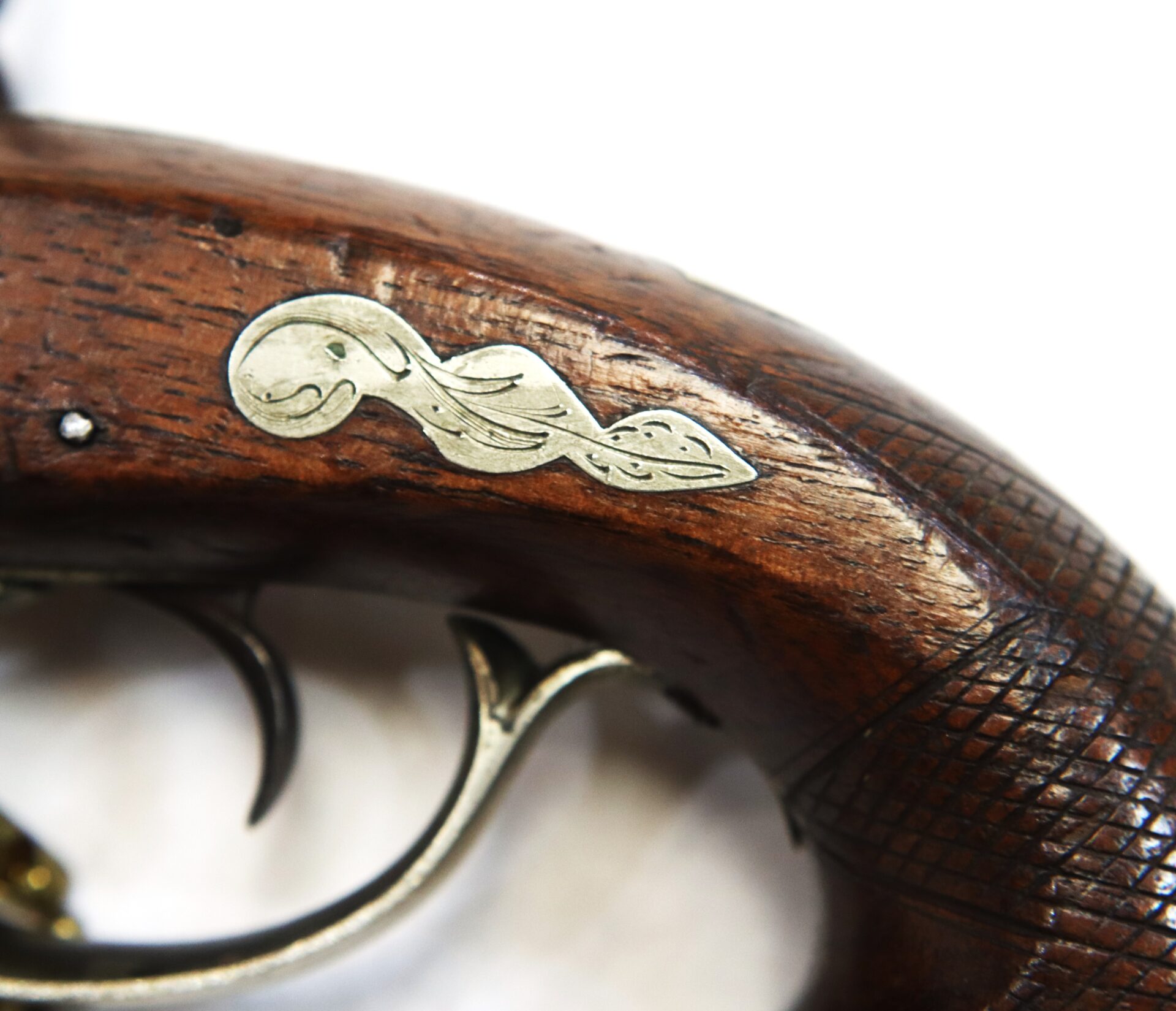 Close-up of a wooden firearm handle with metal inlay featuring an ornate design, and visible checkered texture near the metal trigger.