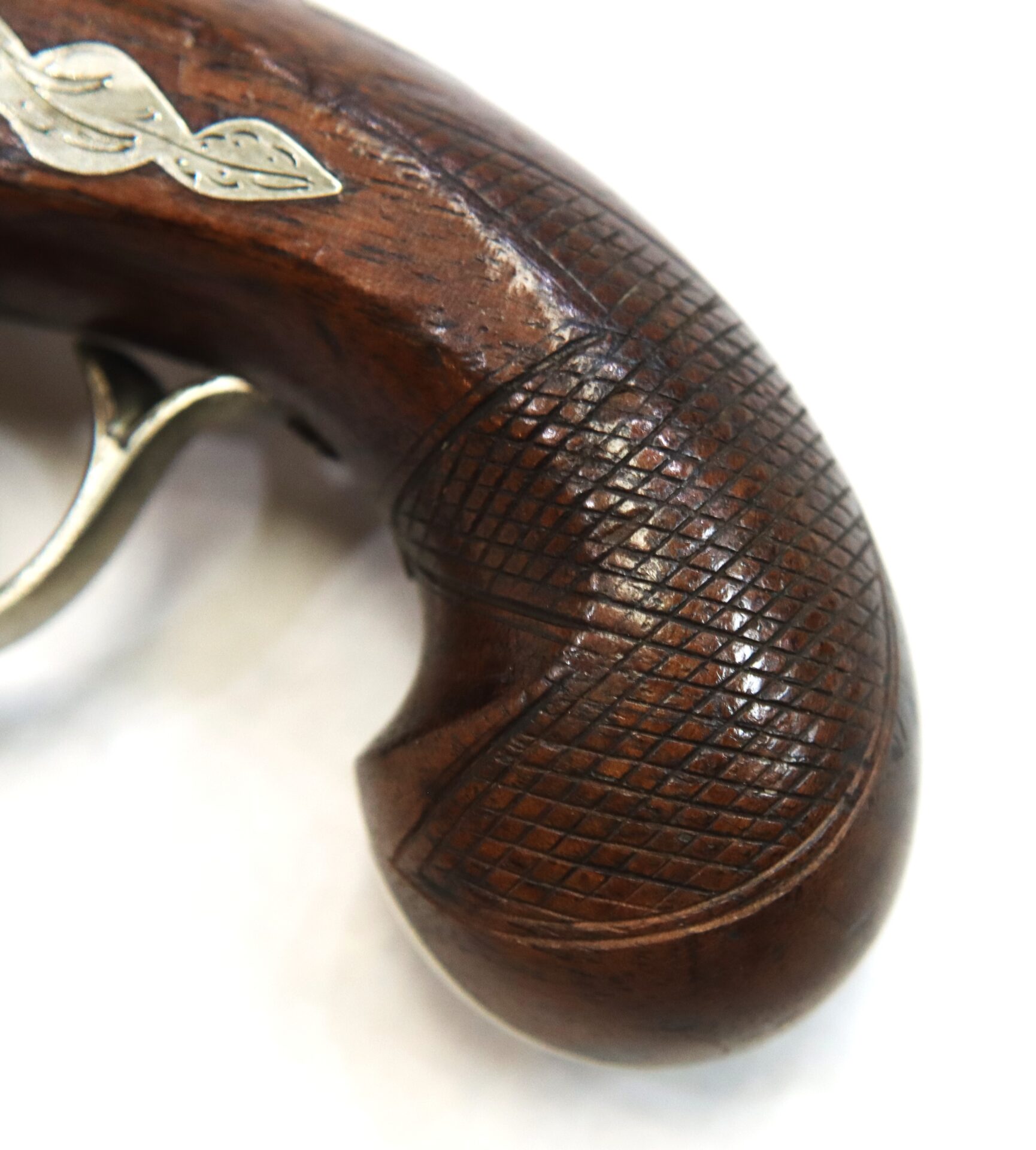 Close-up of a wooden pistol grip with detailed checkered engraving and a decorative metal inlay, against a white background.