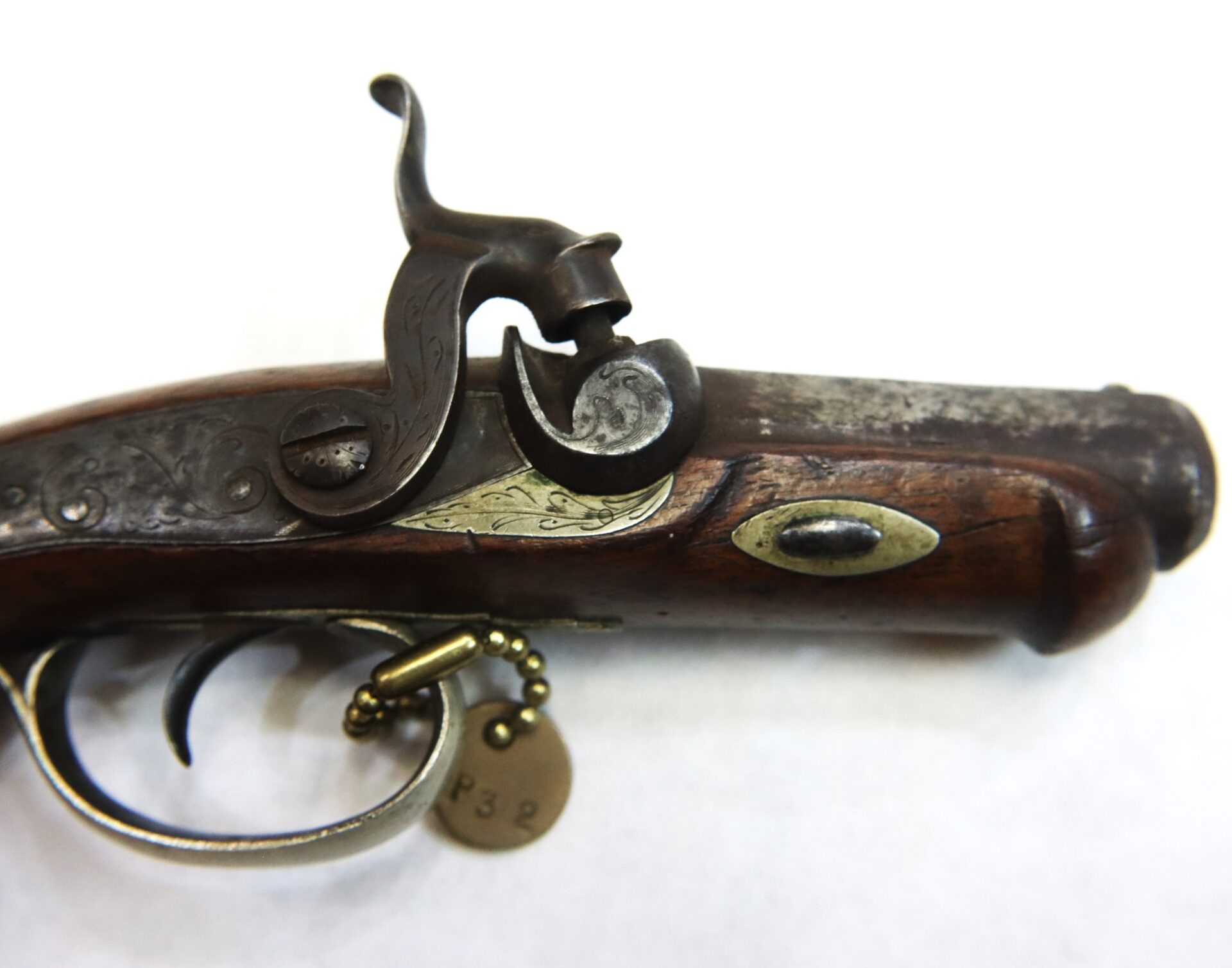 Close-up of an antique flintlock pistol with ornate metalwork on the lock and a numbered tag attached to the trigger guard.