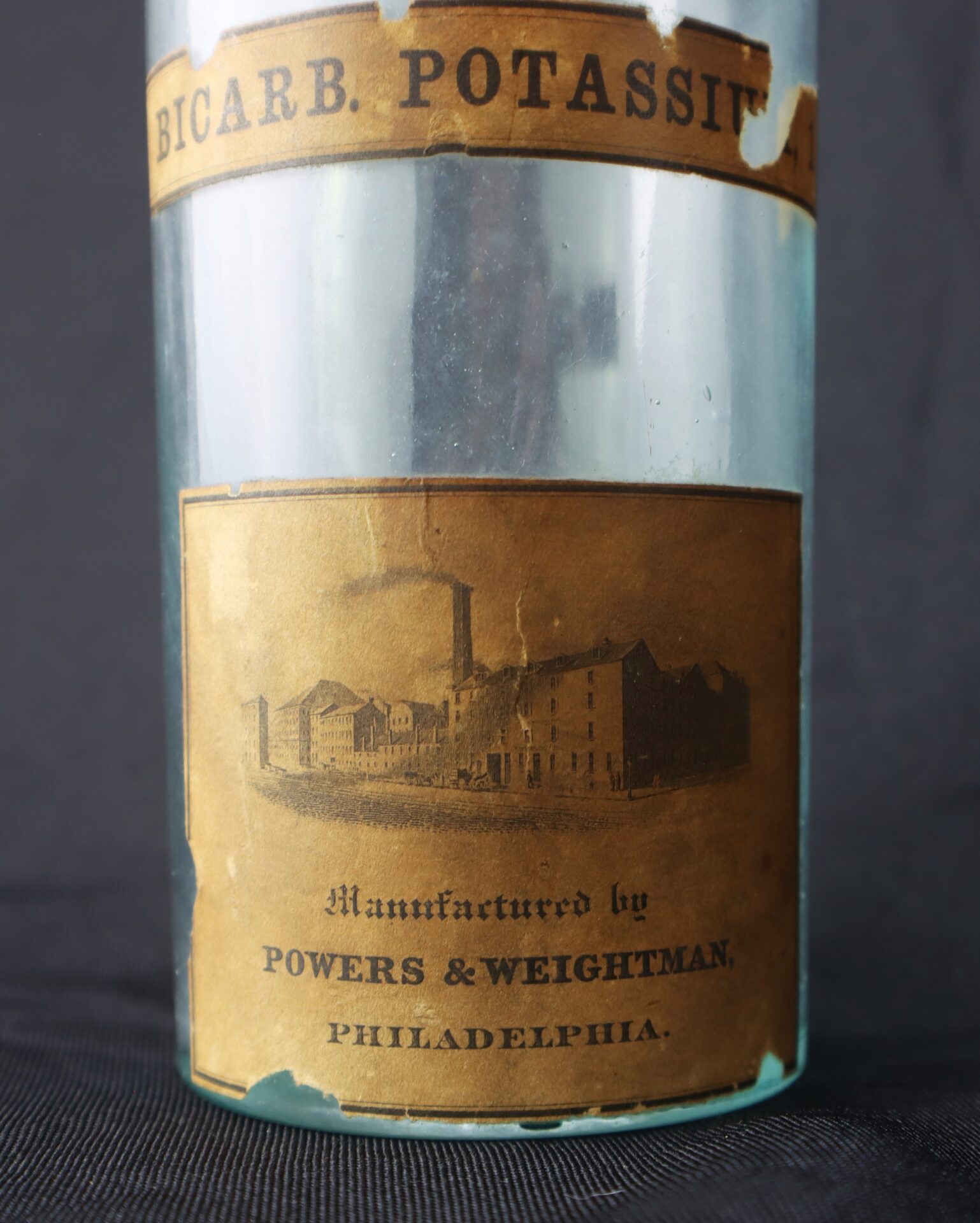 A vintage glass bottle with a brown label reading “BICARB. POTASSII” and “Manufactured by POWERS & WEIGHTMAN. PHILADELPHIA” above an illustration of a factory.