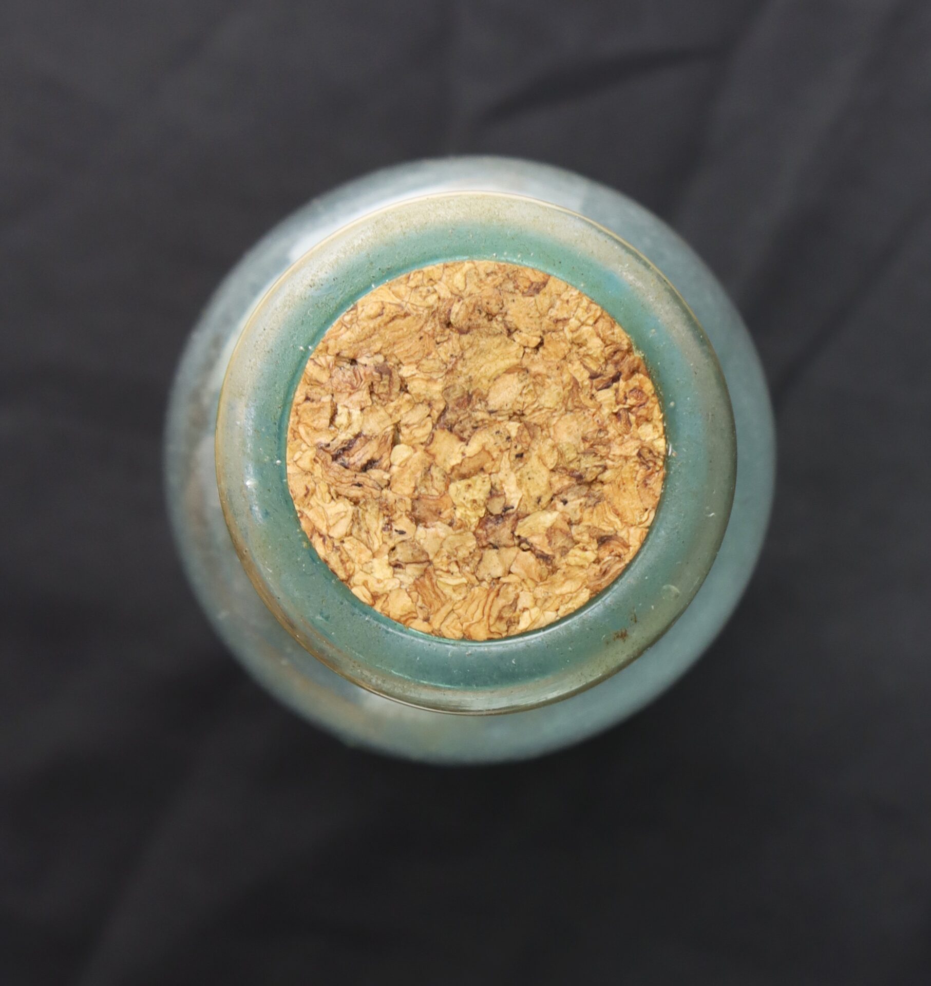 Top view of a round glass bottle with a cork stopper, placed on a dark fabric background.