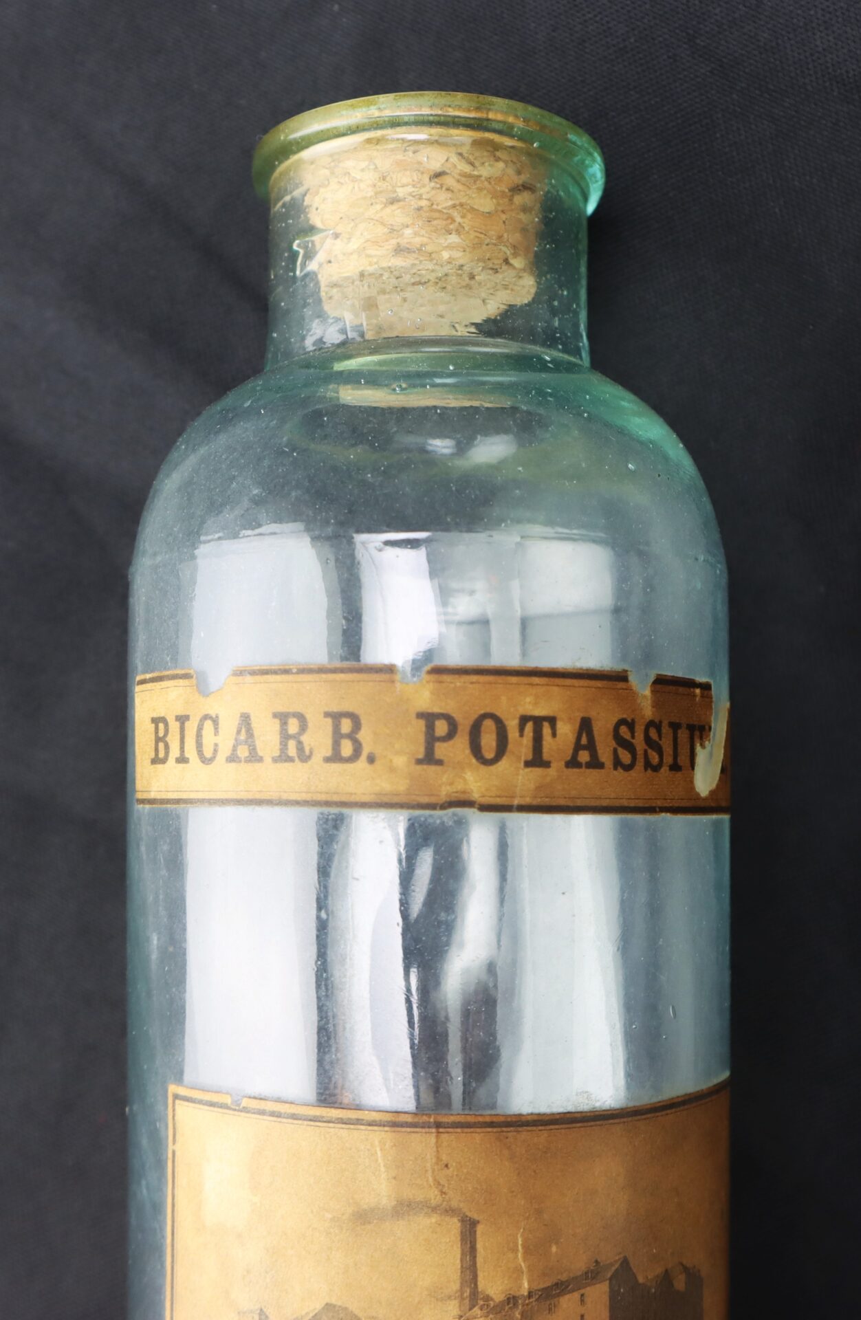 A vintage glass bottle with a cork stopper labeled "BICARB. POTASS." against a dark background.