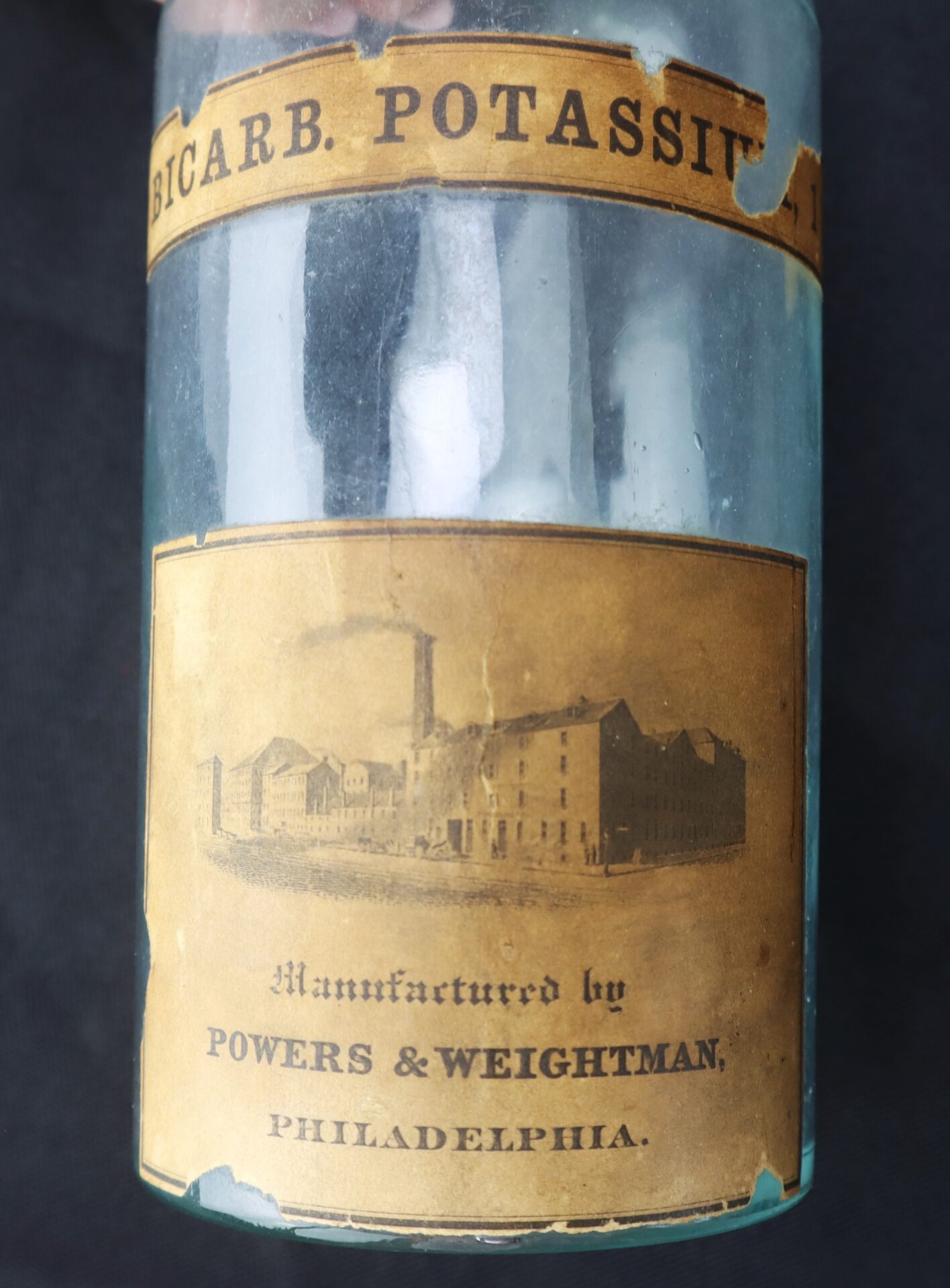 A vintage glass bottle with a worn label reading "BICARB. POTASSII", featuring an illustration of a factory and stating "Manufactured by POWERS & WEIGHTMAN, PHILADELPHIA.