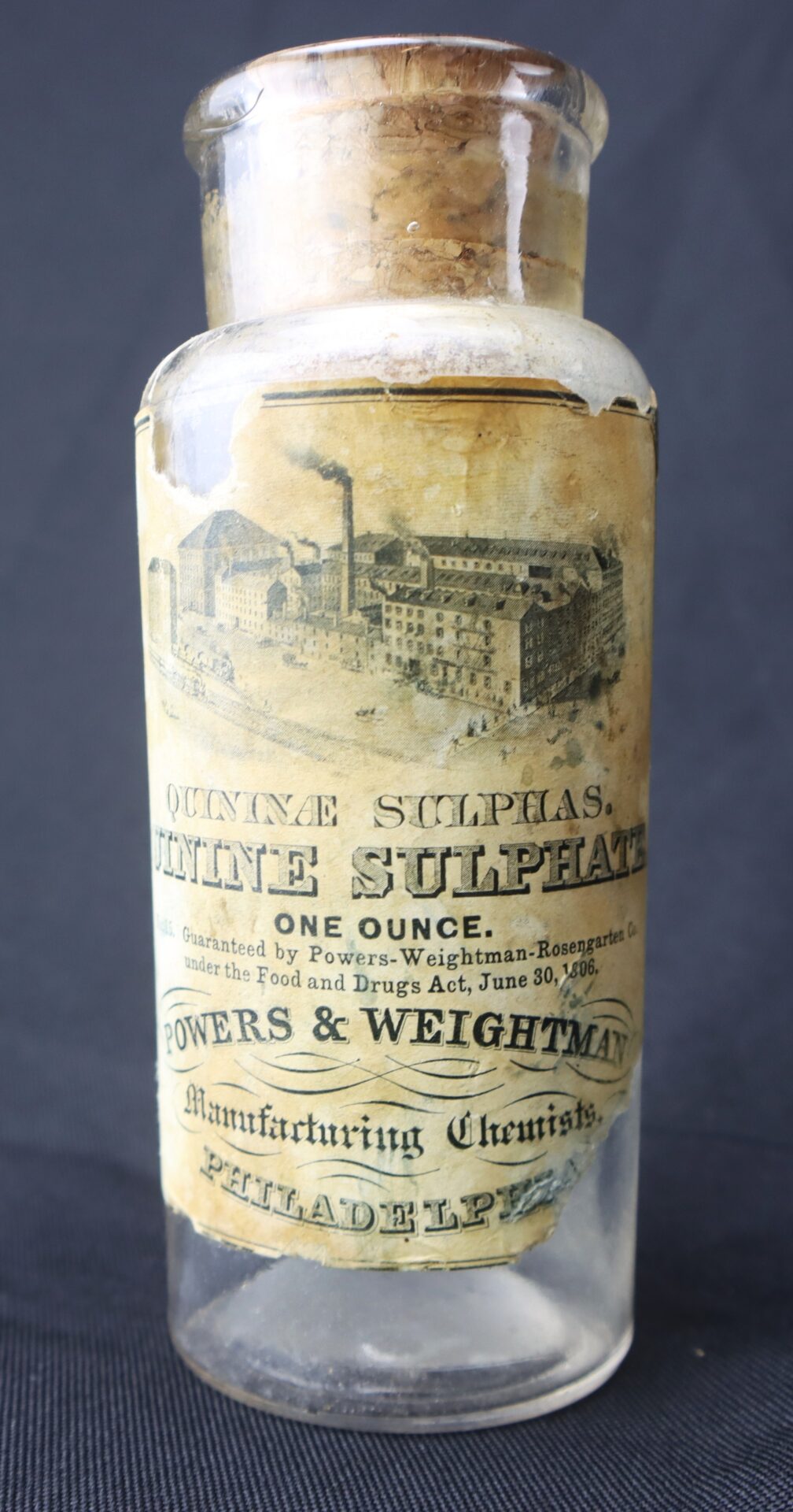 A vintage glass bottle with a cork, labeled "Quinine Sulphate, One Ounce" by Powers & Weightman, Manufacturing Chemists, Philadelphia.