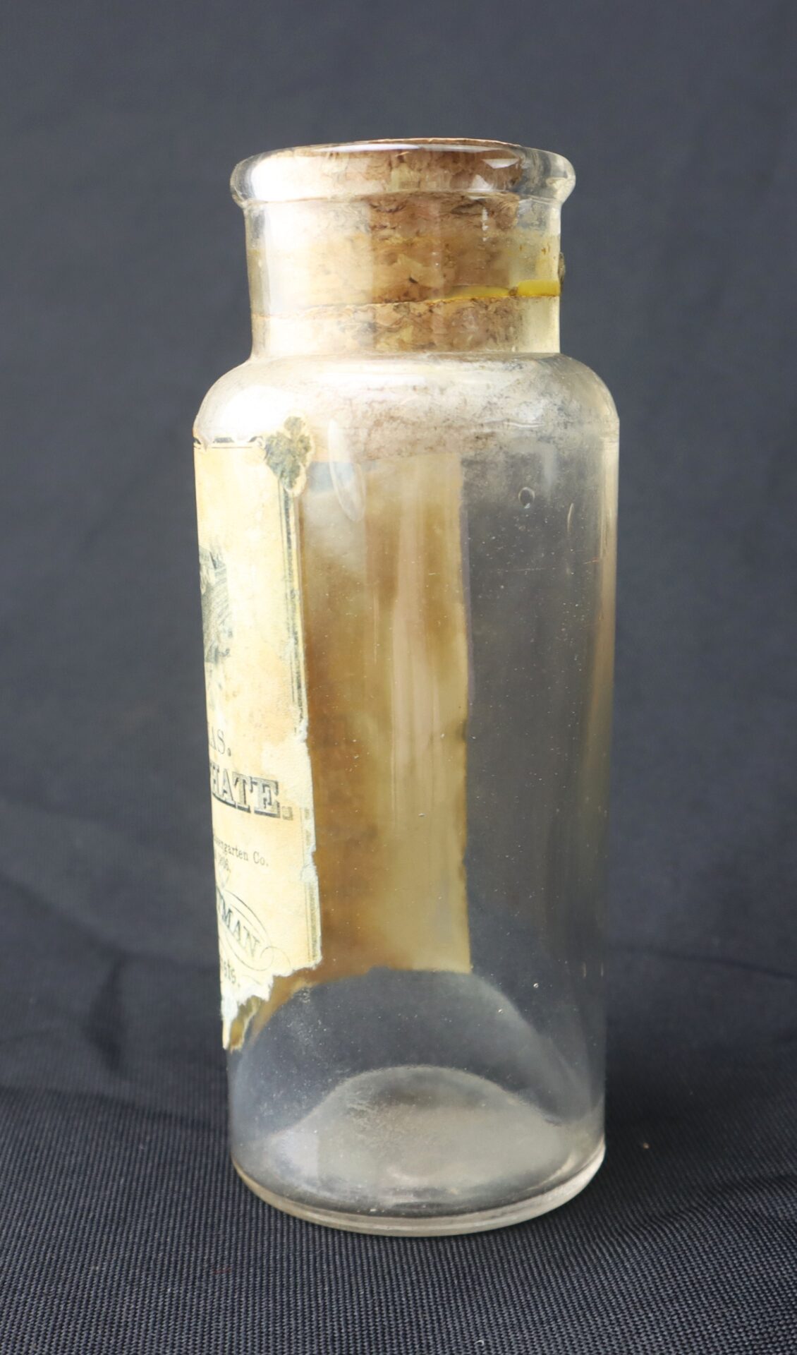 A clear glass bottle with a cork stopper and a partially torn, faded label, standing upright against a dark background.