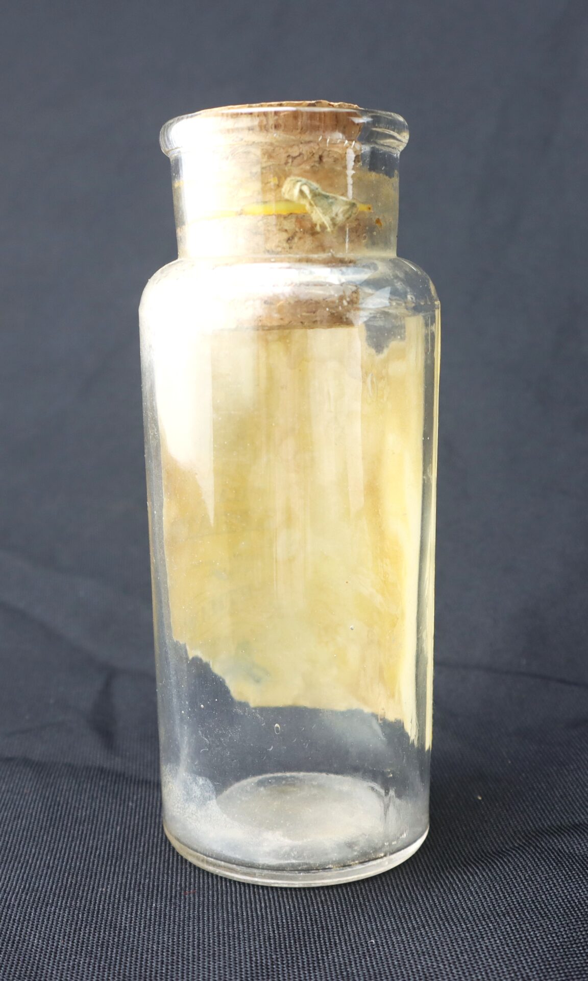 A clear glass jar with a cork lid containing a folded, yellowed piece of paper, set against a dark background.