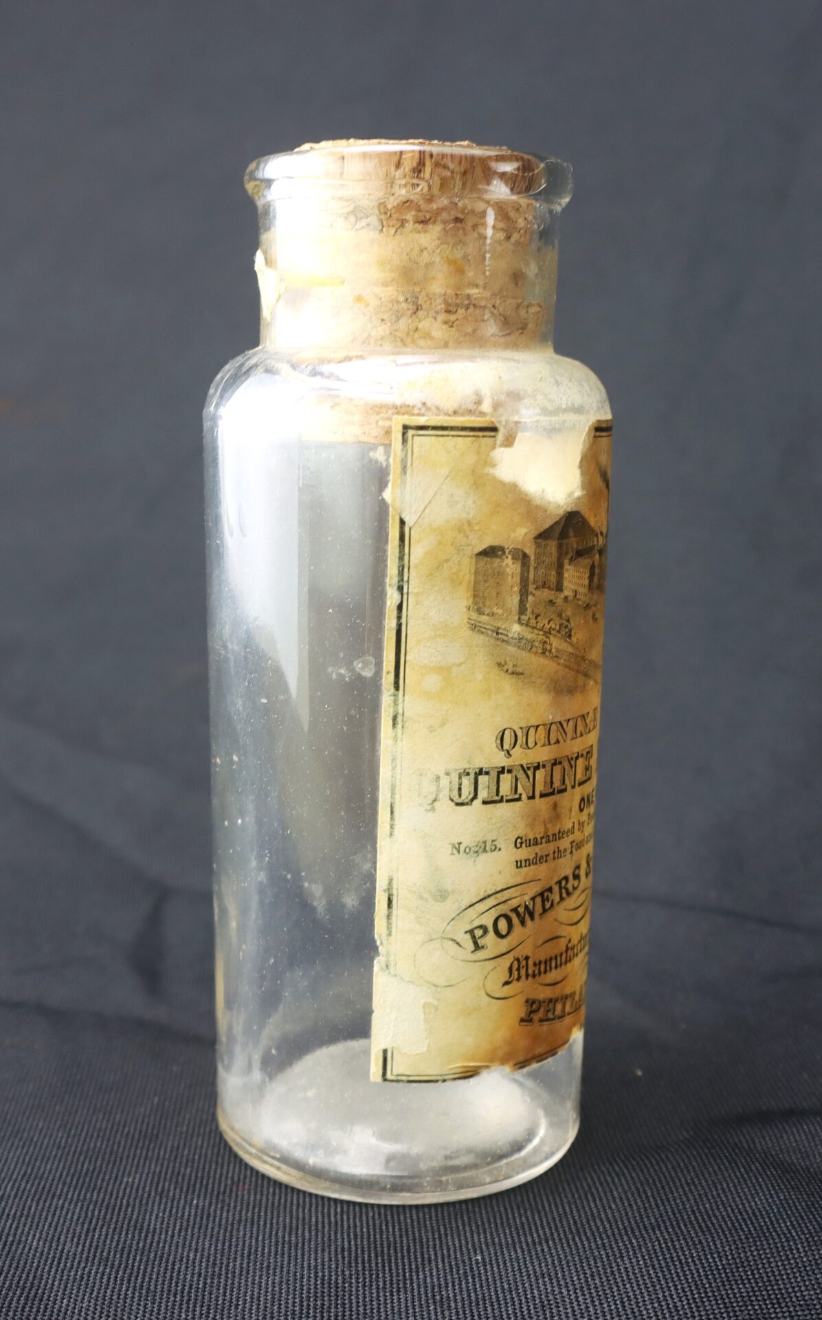 A vintage glass bottle with a cork stopper and a partially torn, yellowed label displaying text and an image, set against a dark background.