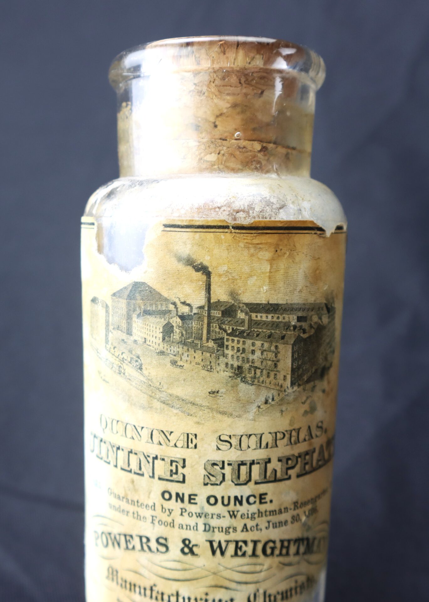 A vintage glass bottle with a cork stopper, labeled “Quinine Sulphate, One Ounce” from Powers & Weightman, featuring an illustration of a factory.