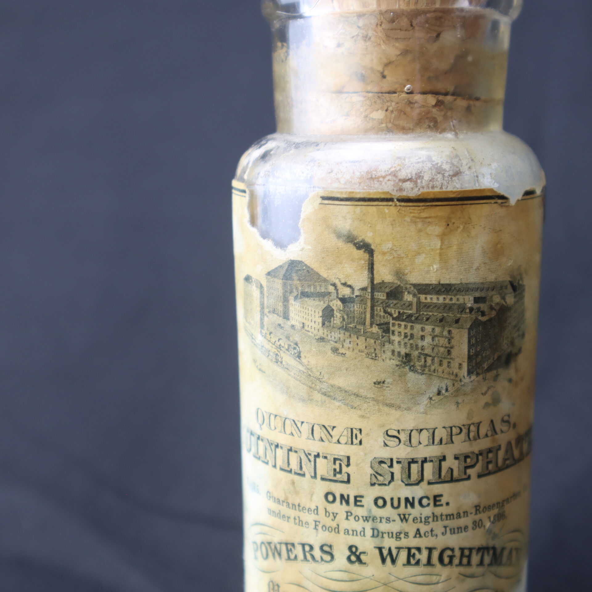 Close-up of an old glass bottle with a worn label reading "Quinine Sulphate, One Ounce, Powers & Weightman," featuring an illustration of a factory.