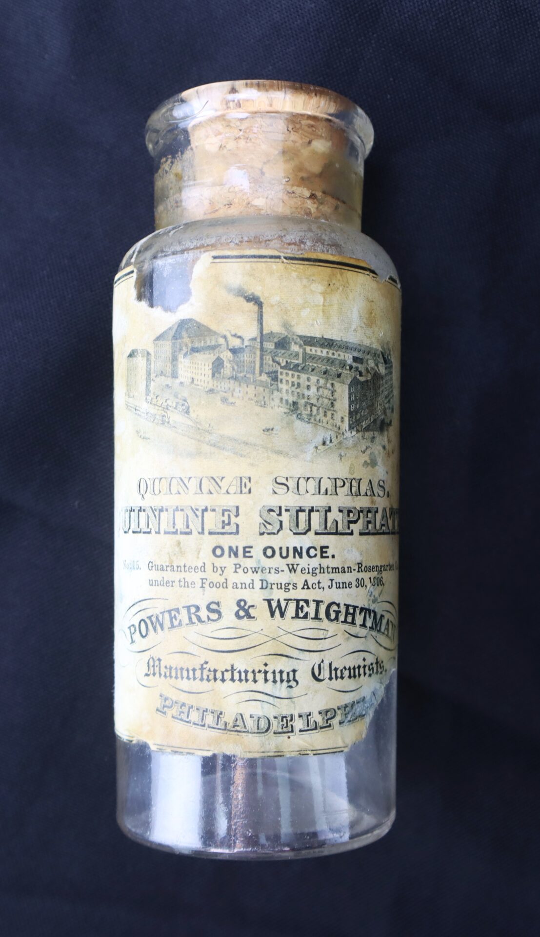 A vintage glass bottle labeled "Quinine Sulphate, One Ounce" with a cork stopper and worn paper label from Powers & Weightman, Manufacturing Chemists, Philadelphia.