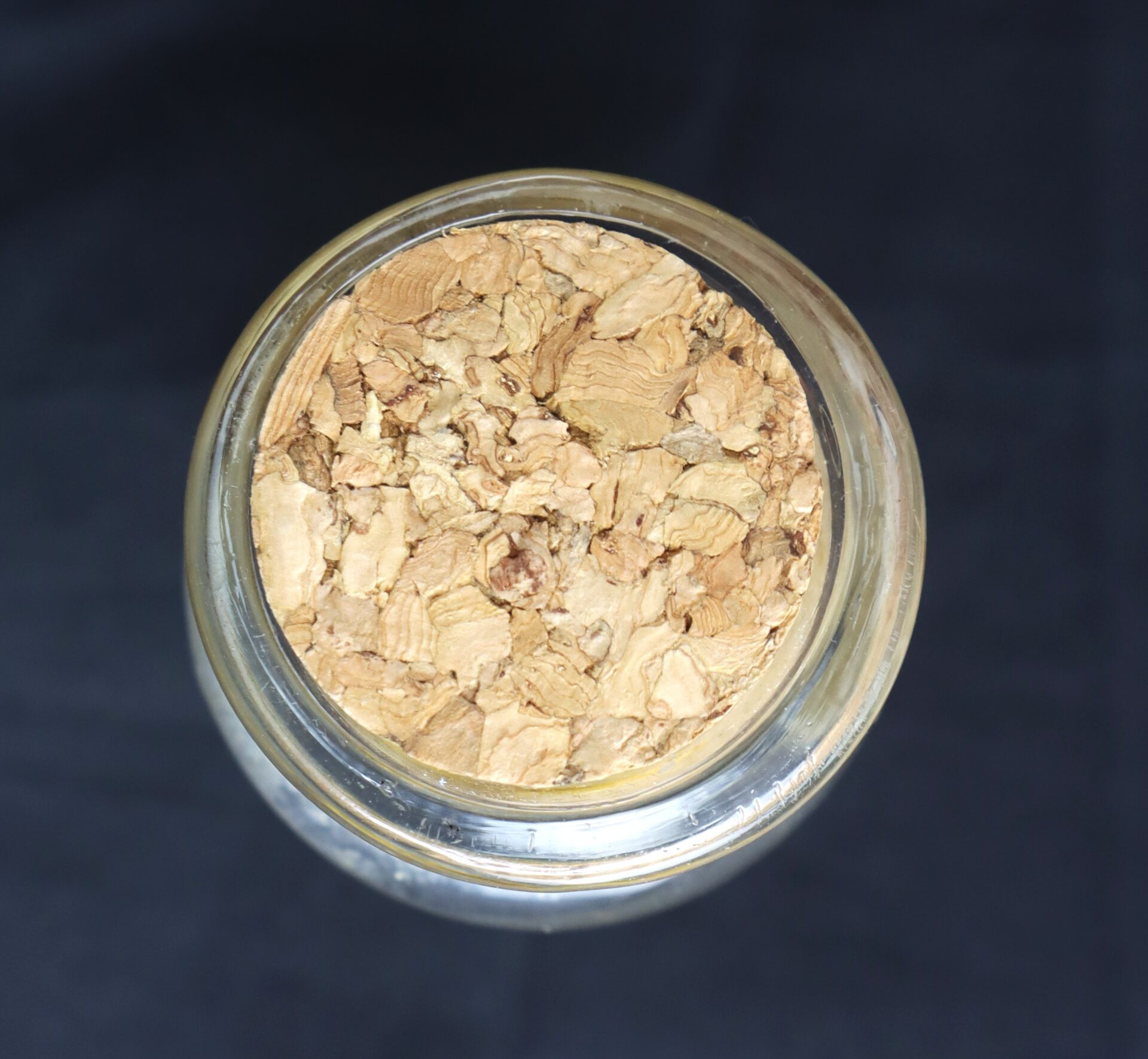 A glass jar filled with dried, light brown plant material, viewed from above against a dark background.