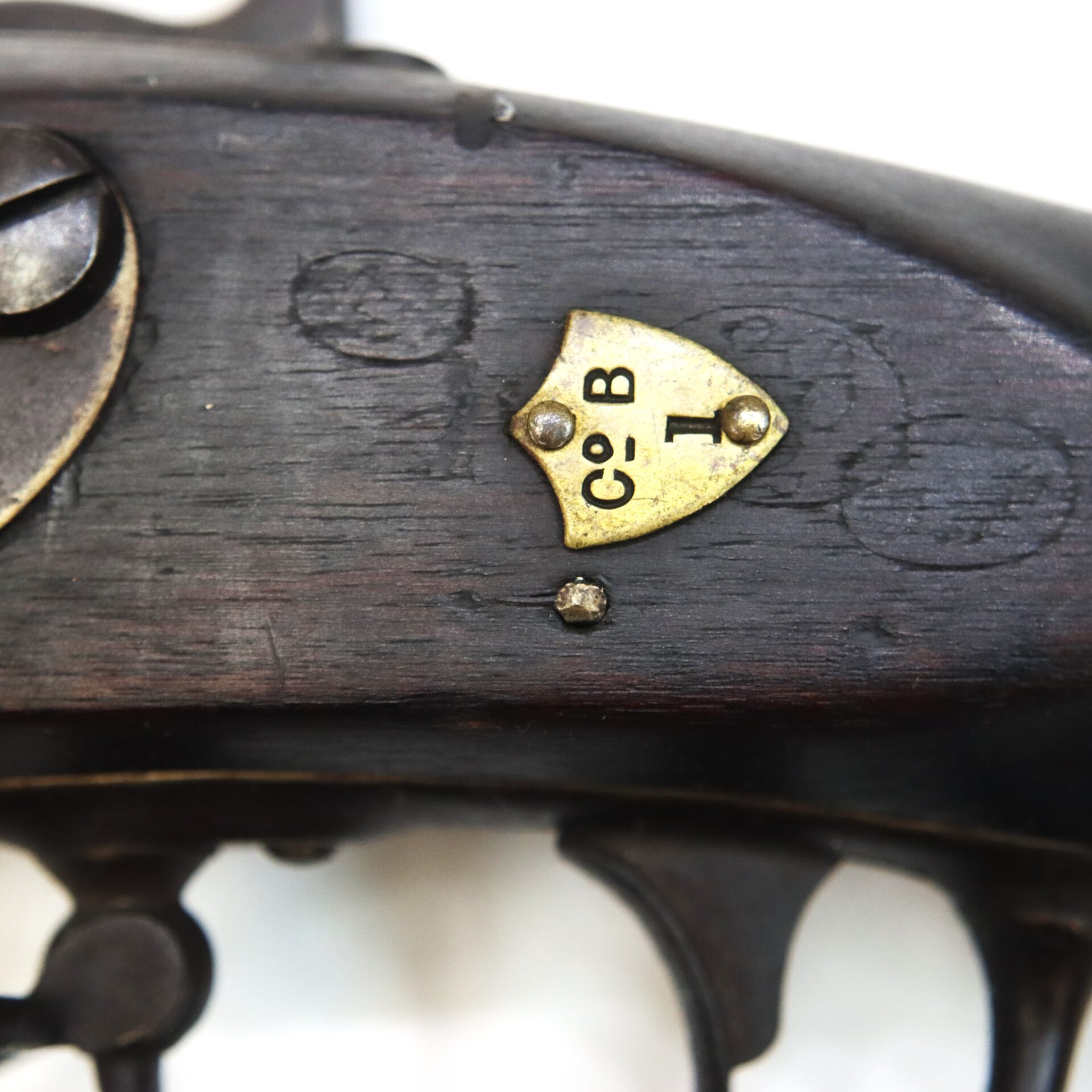 Close-up of a dark wooden firearm stock featuring a small brass plate inscribed with "C•B" and a symbol, secured with a single screw.