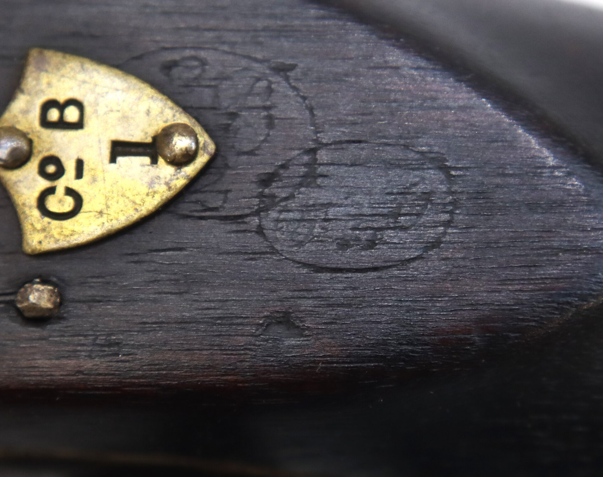 Close-up of a dark wooden surface with a brass plate labeled "Co. B 1" attached by three screws; faint circular markings are visible on the wood.